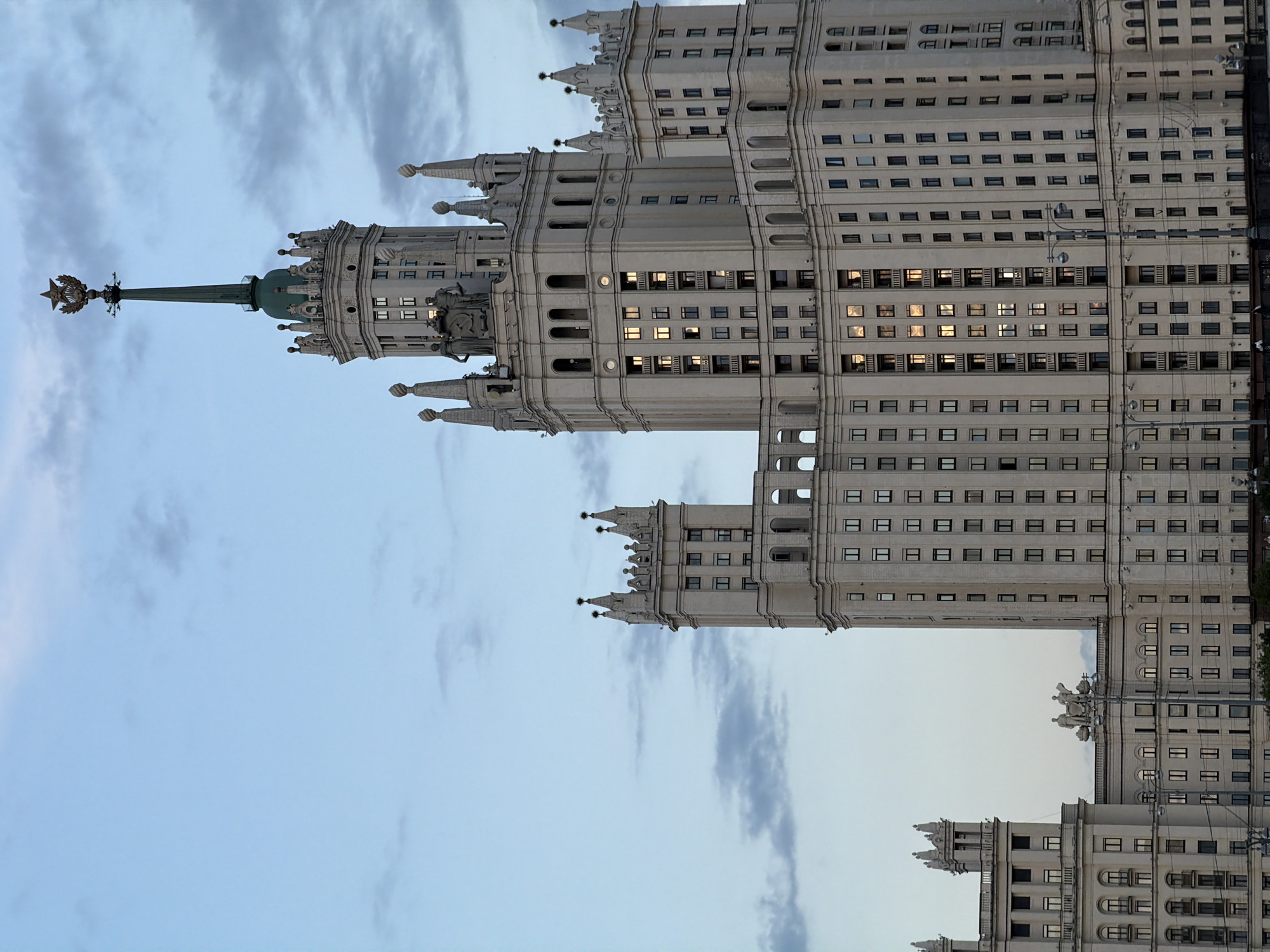 The image displays a towering, ornate building against a cloudy sky, oriented vertically (when rotated 90 degrees counter-clockwise). The architectural style is characteristic of Stalinist Empire architecture, featuring a monumental scale, tiered structure, and extensive decorative elements. The central part of the building is crowned with a tall, slender spire that culminates in a gold-colored star and wreath motif.
The building's facade is light-colored, likely stone or stucco, and is punctuated by numerous windows. Many windows are rectangular, while some in the more decorative lower sections are arched. Several windows reflect a warm, golden light, possibly from interior lighting or the setting sun. Ornate details such as smaller spires, turrets, decorative ledges, and what appear to be sculptural groups are visible across different sections of the building.
The sky is a soft blend of light blue and grey, with diffused clouds, suggesting an overcast day or twilight hours. No human activity or specific objects other than the building and sky are visible. Based on its distinctive architectural features, including the prominent spire and star, the structure is identifiable as one of Moscow's Stalinist skyscrapers, commonly known as the "Seven Sisters," located in Moscow, Russia. There is no visible text in the image.