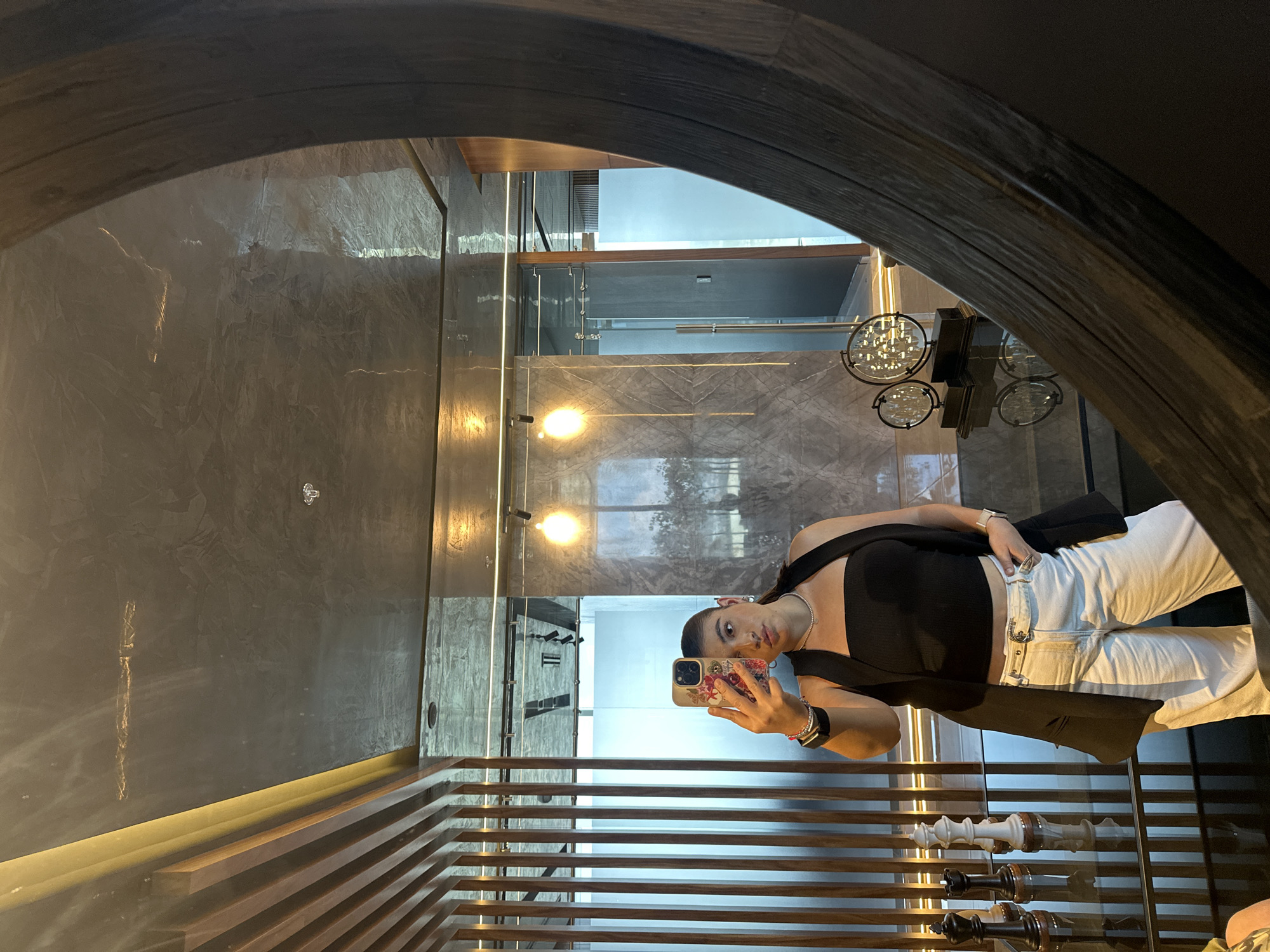 The image shows a woman taking a selfie in front of a mirror. She is wearing a black top, white pants, and a black blazer. She is holding a phone with a floral case. The background of the image shows a modern interior with marble walls, a wooden slat ceiling with integrated lighting, and glass dividers. In the reflection of the mirror, there are decorative light fixtures and a glimpse of an upper floor. The location is San Bernardino Tlaxcalancingo, Mexico.