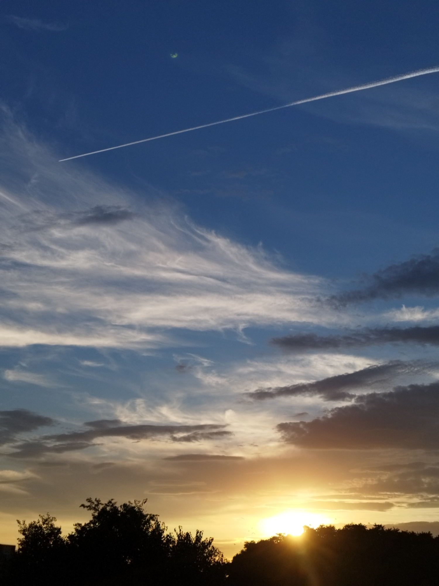 A low-angle, vertical, full shot shows a sunset over Bucharest, Romania. A bright yellow sun peeks over a dark silhouetted tree line, casting rays into a sky filled with layers of clouds ranging from white to dark gray. Wispy white clouds are scattered across the upper part of the sky, with a contrail from an airplane cutting diagonally from the upper left to the middle right. In the far upper left corner of the sky, a small, blurry green circular object is visible, possibly a distant light or lens flare. The trees are dense and dark, obscuring any ground detail, and appear to be a mix of deciduous and evergreen types. The overall atmosphere is one of a colorful and dramatic end to the day.