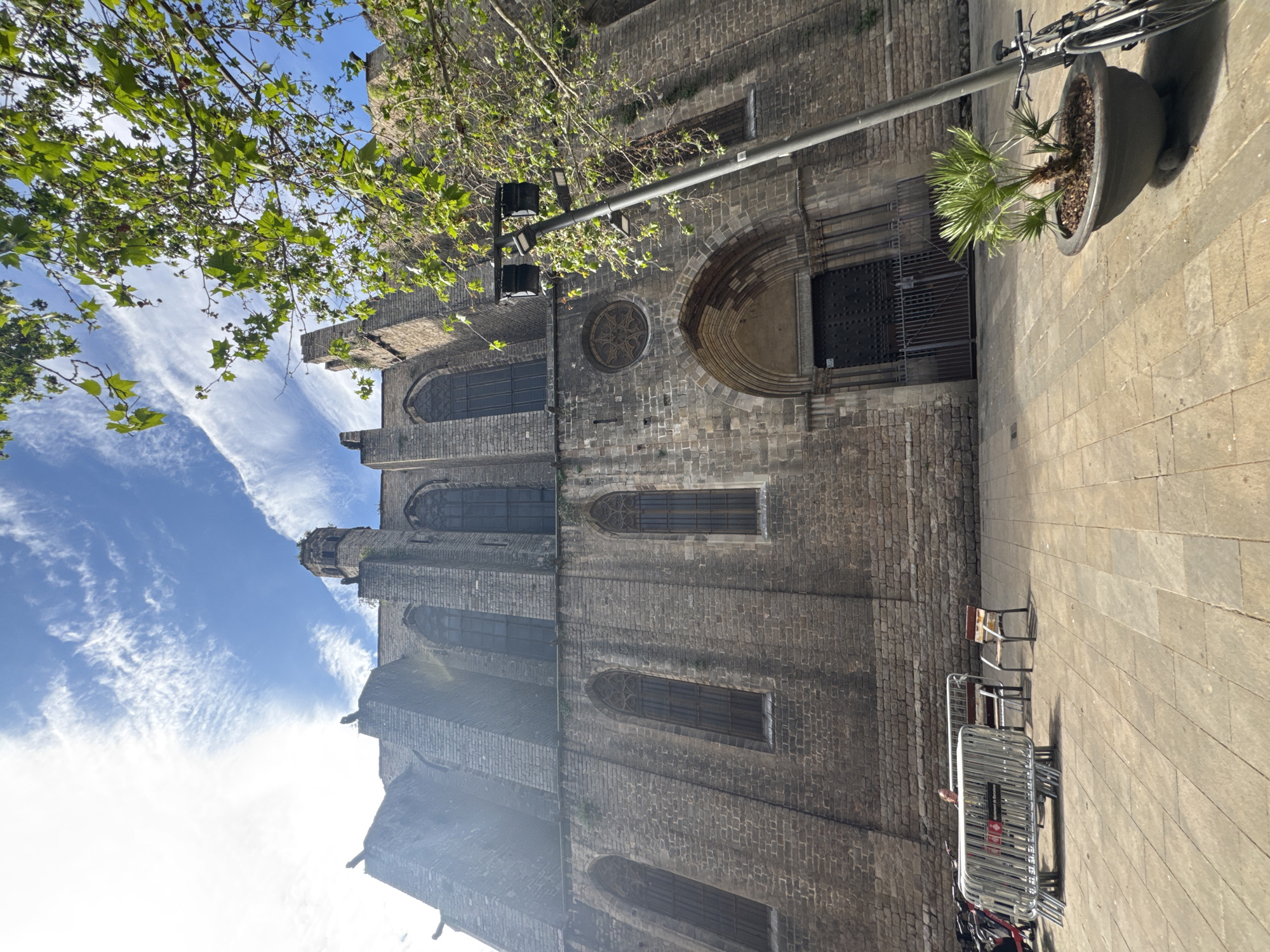 The image displays an exterior view of a large, historic stone building, likely a church or cathedral, in an urban setting identified as Barcelona, Spain. The building is constructed from dark, textured stone blocks and features several tall, narrow arched windows with grilles. A prominent circular window with a decorative star-like tracery is situated above a large, deep-set arched entrance, which is secured by a heavy metal gate. Architectural details, including what appear to be gargoyles, are visible on the upper sections of the structure.
In the foreground and to the right, a paved street or sidewalk is visible, composed of both smaller cobblestones and larger, lighter flagstones. A grey street lamp pole extends across the mid-ground. Beneath it, a large dark planter holds a small, vibrant green palm-like plant. A bicycle is parked nearby. Further right, a stack of metallic crowd barriers and a single chair with a wooden seat are positioned on the sidewalk.
Overhead, a tree with bright green leaves partially frames the upper left corner of the scene, its branches extending into the blue sky. The sky is clear with scattered, wispy white clouds. The bright daylight and visible shadows suggest a sunny day, likely mid-morning or afternoon. No explicit human activity is depicted. No clearly legible text is visible on any objects within the frame.