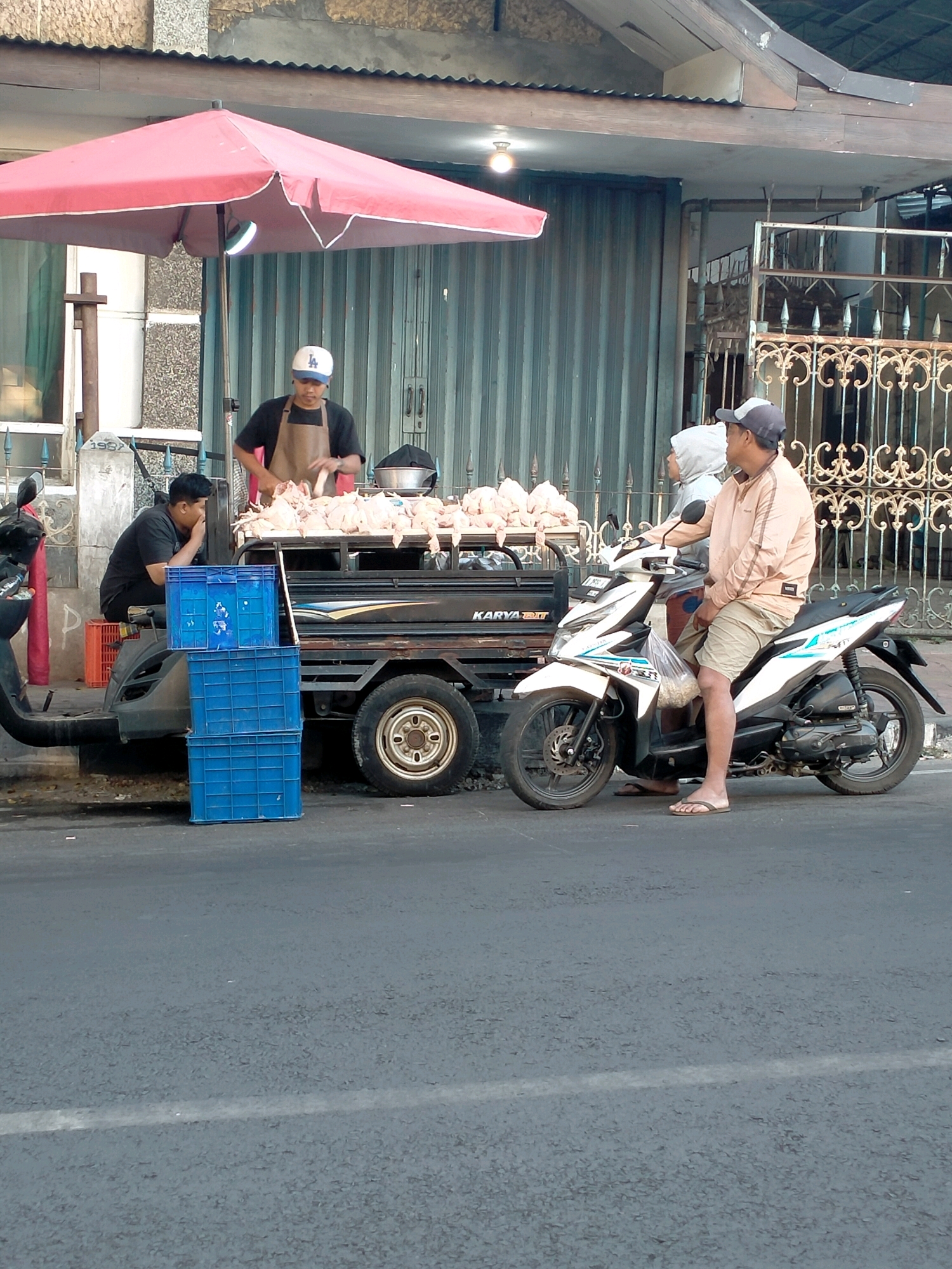 In the city of Batu, Indonesia, an outdoor scene unfolds with a food stall featuring a display of whole chickens. A man wearing a brown apron and a white baseball cap, identified as a vendor, is actively preparing the poultry. Beside him, on a trailer that appears to be attached to a vehicle, a large quantity of dressed chickens are arranged. To the left of the vendor, another man sits on a red crate, observing the proceedings. In the foreground, two individuals are seated on a motorcycle. One, wearing a light-colored jacket and shorts, is looking towards the vendor, while the other, obscured by the first, is looking away. The environment suggests a street setting with a building in the background featuring a metal roll-up door and an ornate metal fence. Several blue plastic crates are stacked near the trailer.