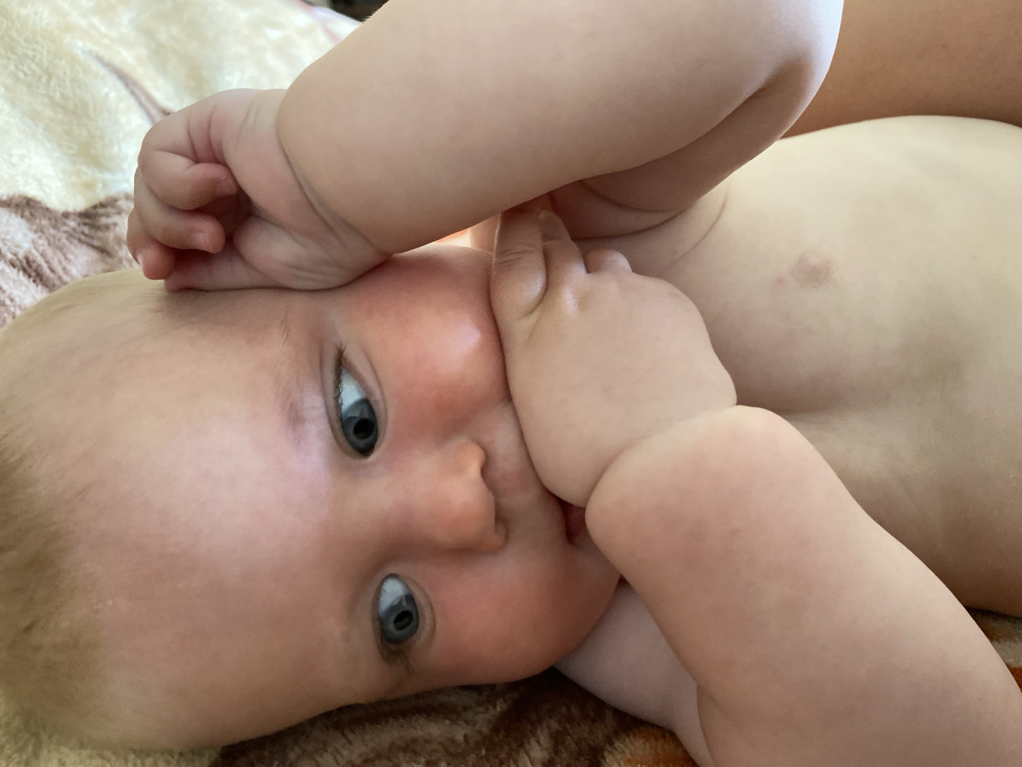 A close-up shot shows a baby lying on its back on a patterned blanket. The baby's head is tilted to the right, and its eyes are open, looking towards the camera. One of the baby's hands is placed over its mouth, and the other is bent over its head. The baby's skin is rosy, and it has blonde hair. The background is a soft, blurred blanket with a brown and beige pattern.