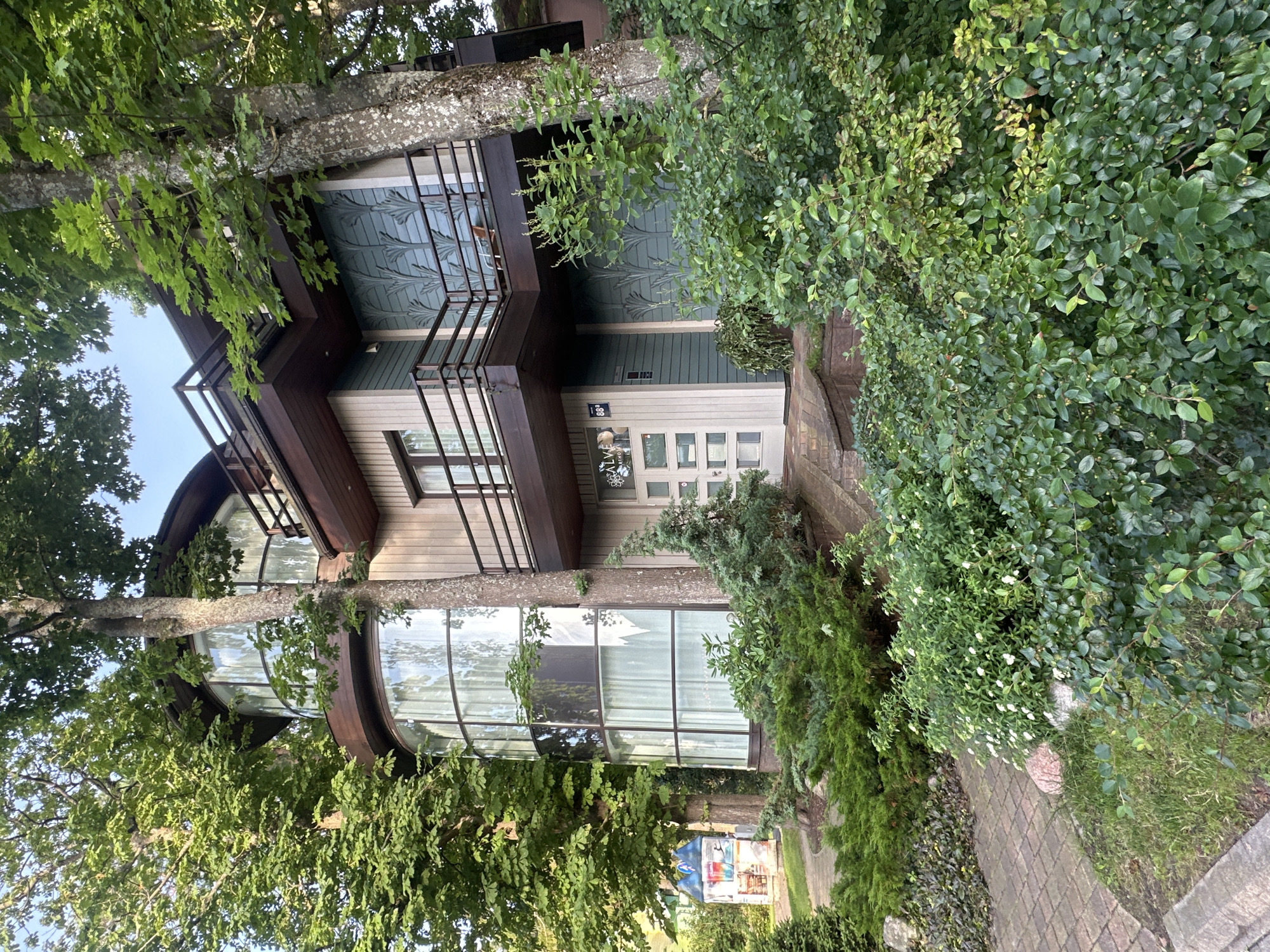 The image shows a modern multi-story house with large curved windows and dark brown balconies, surrounded by lush green trees and bushes. The house has light-colored siding and decorative blue panels on the upper levels. A brick pathway leads to the entrance, which features a white door with glass panels and a sign that reads "SALVE APARTMENT." The property is located in Jurmala, Latvia, a resort city on the Baltic Sea known for its sandy beaches and pine forests. The overall impression is one of a serene and well-maintained residential property nestled within a natural setting.