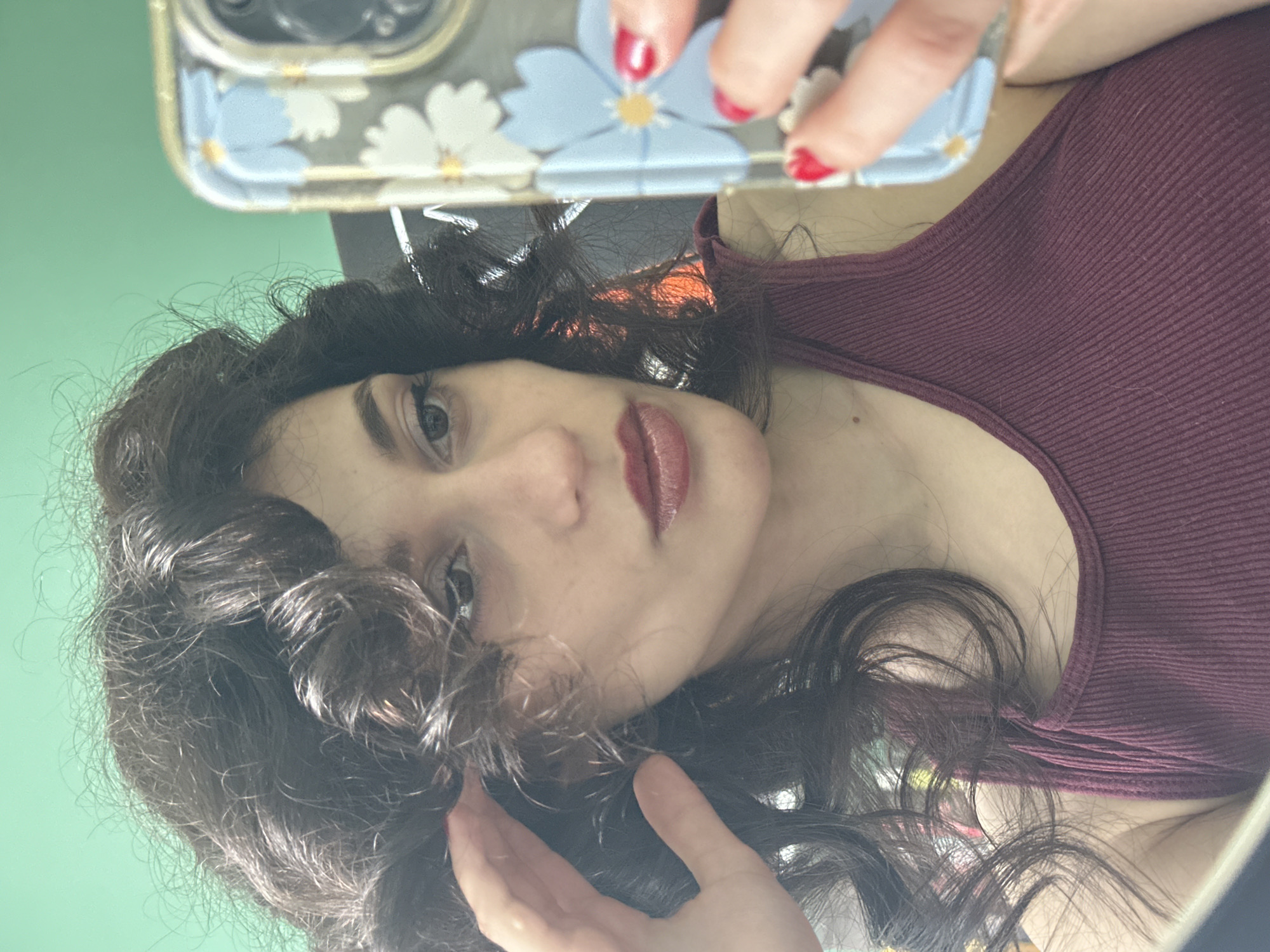 This is a portrait-oriented selfie of a young woman in Sofia, Bulgaria. She has dark, curly hair that frames her face, and she is wearing a deep red, ribbed tank top. Her makeup includes dark eyeliner and a berry-toned lipstick with a glossy finish. One hand, with bright red nail polish, is visible near the top of the frame, touching a phone case decorated with blue and white flowers. The background is a solid, light green wall. The lighting suggests it could be daytime, possibly indoors given the controlled environment. The woman's gaze is directed towards the camera, and her expression appears neutral or perhaps slightly thoughtful. There is no visible text in the image.
