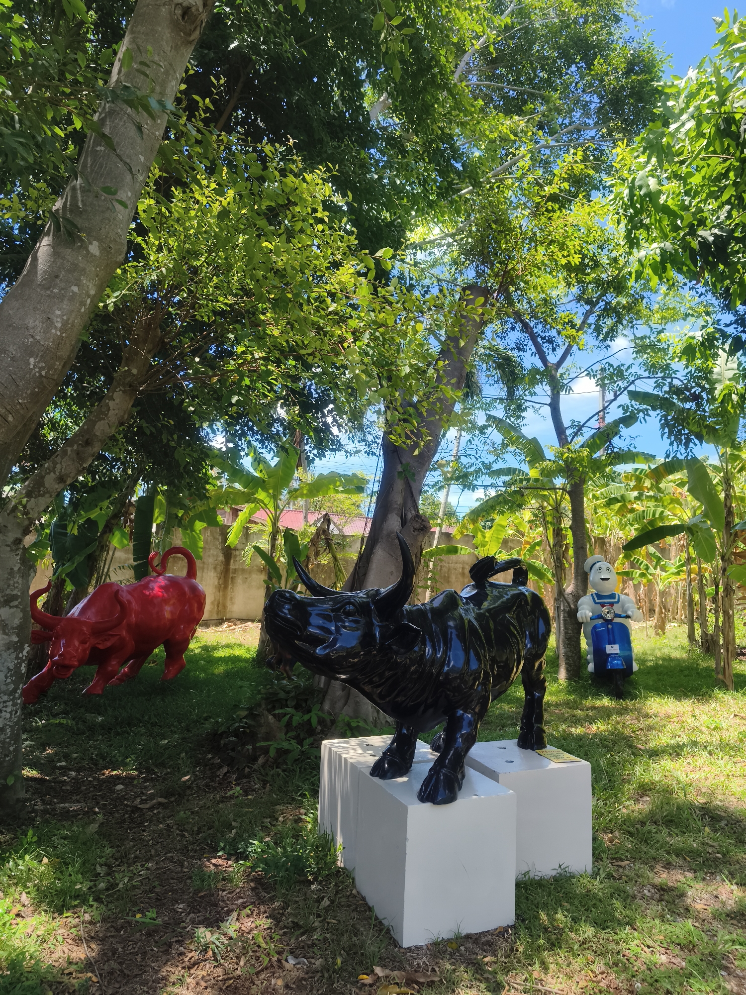 The image shows an outdoor art installation in Baan Bo Phut, Thailand. A red bull statue stands on the left, partially obscured by a tree. In the center, a black water buffalo statue is elevated on a white rectangular platform. To the right, a white and blue statue resembling the Stay Puft Marshmallow Man from Ghostbusters is depicted riding a blue scooter. The background includes green trees and banana plants, with a blue sky visible through the foliage. The ground is covered in grass and scattered leaves.