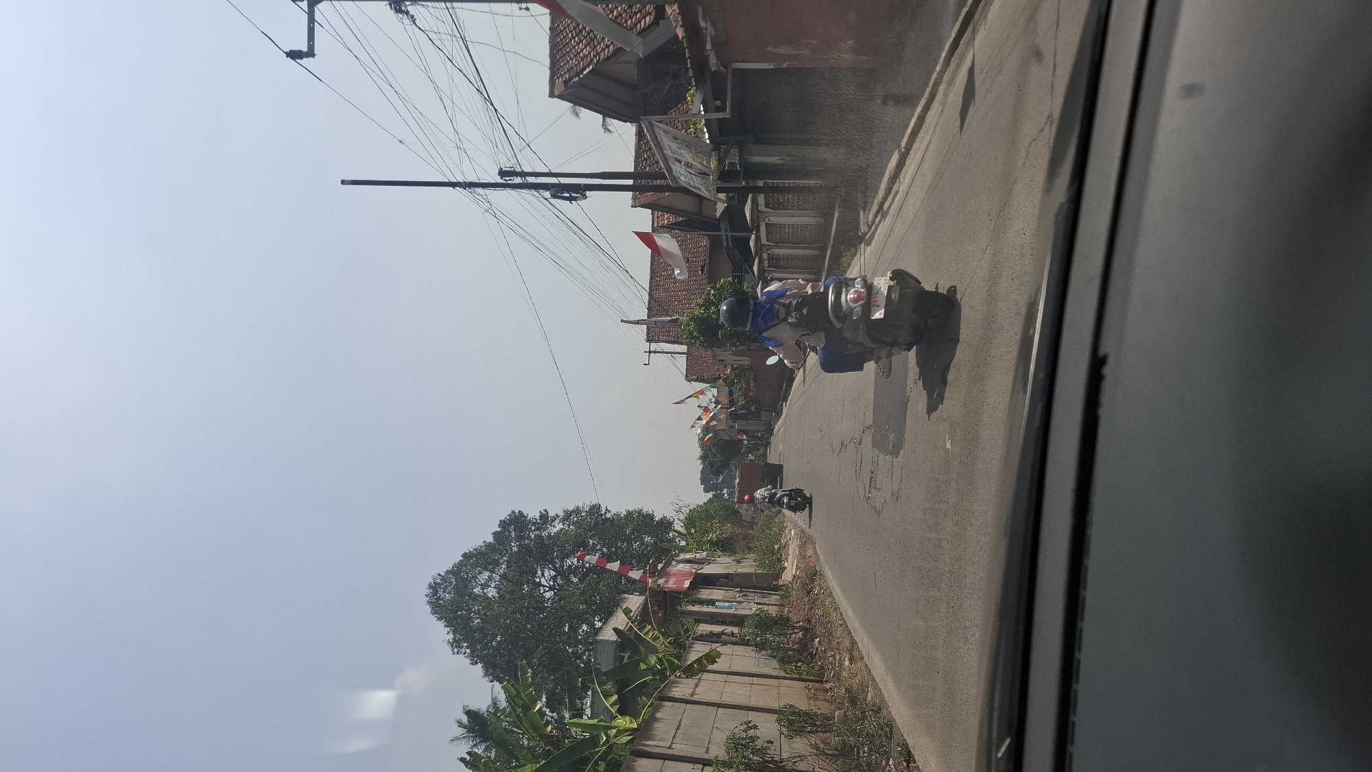 The image depicts a street scene in Sirnagalih, Indonesia, on a bright, clear day. The perspective is from inside a vehicle, looking forward through the windshield. A paved road, showing some cracks and wear, stretches into the distance, flanked by houses and a concrete wall on the left. Several Indonesian flags, red and white, are displayed on poles and attached to buildings, suggesting a national holiday or celebration.
Two individuals are riding motorcycles on the road. The closest motorcycle, in the foreground, is ridden by a person wearing a blue headscarf and a backpack, with a dark helmet. They are moving away from the camera. Further down the road, another motorcycle with a rider wearing a red helmet is also traveling in the same direction. In the far distance, a truck is visible.
The environment is characterized by residential buildings with tiled roofs, some with visible decorations. There are also trees and vegetation, including banana plants, along the roadside. Overhead, power lines are strung between utility poles, a common sight in urban and semi-urban areas. The sky is pale blue and hazy, indicating warm weather conditions. The overall impression is of a typical day in a populated area of Sirnagalih.