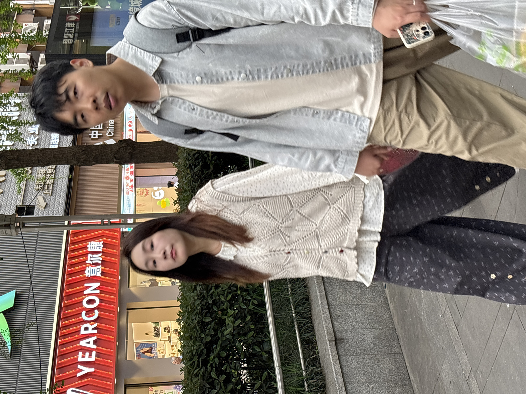 A slightly tilted medium outdoor shot presents a young man and woman on a city street. The man, positioned in the top half of the image, has short black hair and wears a light grey denim jacket over a white t-shirt and tan trousers. He is holding a plastic bag in his right hand and a smartphone in his left.
The woman, in the bottom half of the image, has long brown hair and wears a white textured cardigan over a light-colored top and patterned black pants. Both appear to be walking.
Behind the woman is a green hedge. To the left is a building with a red sign in Chinese characters: "YEARCON 意尔康" running vertically. Other signs nearby read "China". The presence of this signage helps to determine that the location is likely in the city of Jiangjin, China. A pedestrian walkway runs along the side of the street.