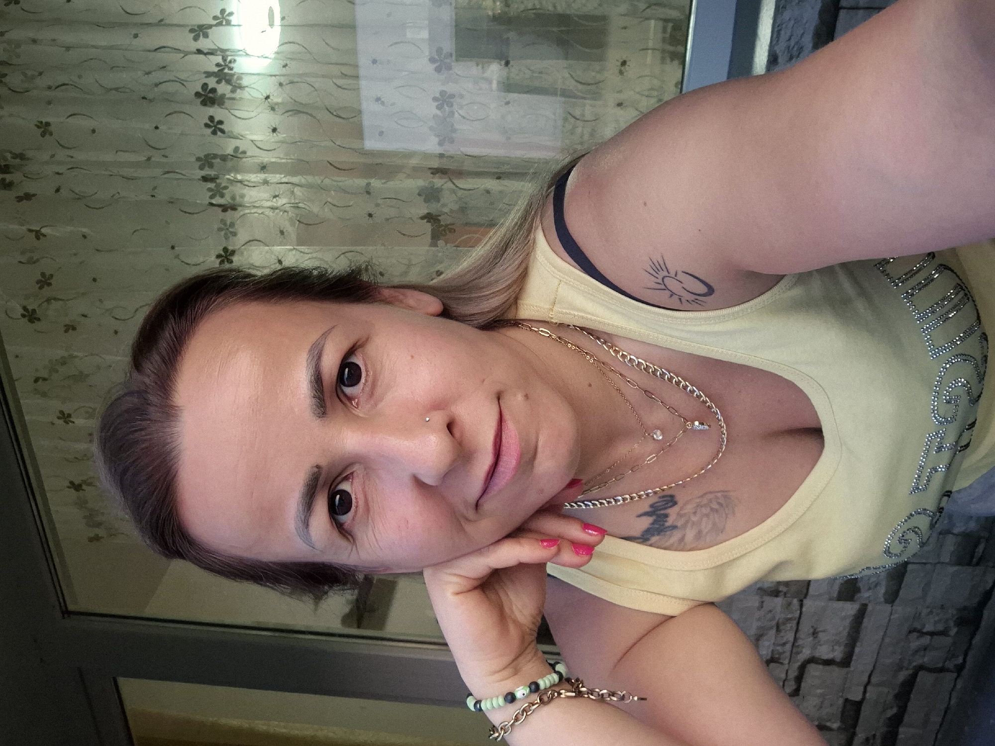 A medium shot shows a woman with brown hair and brown eyes, leaning her head on her hand. She is wearing a light yellow tank top with layered gold necklaces and a bracelet on her left wrist. She has a tattoo on her left arm of a sun and a crescent moon, and another tattoo on her chest. The background appears to be a window with a floral curtain.