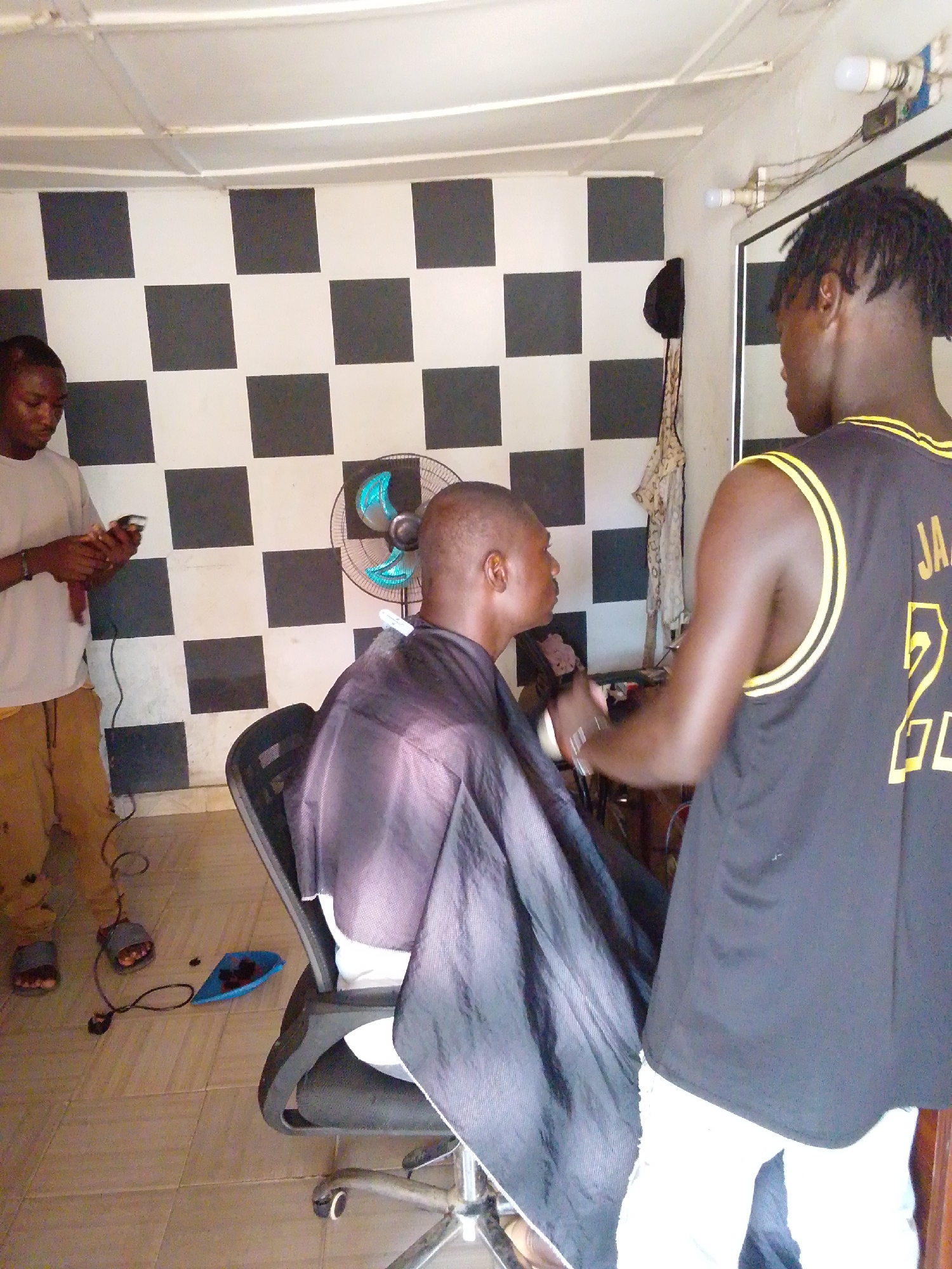 Inside a barbershop in an unspecified city, a man with dreadlocks, wearing a black and gold jersey with "JA" and the number "2" on the back, is giving a haircut to a seated customer draped in a black barber cape. Another man stands in the background, holding a hair clipper. The wall behind them is decorated with alternating black and white squares. A small blue dustpan filled with hair clippings is visible on the tiled floor. There is an electric fan behind the client.