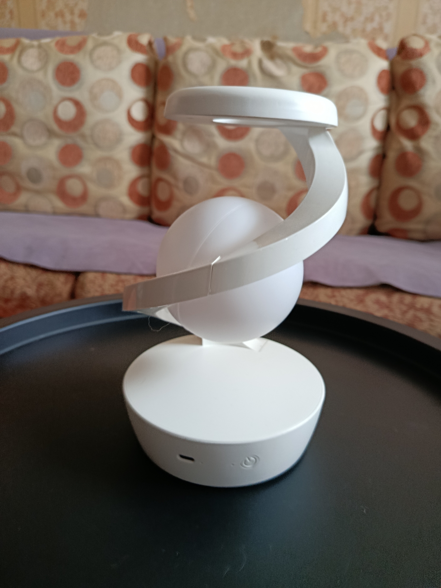 A white, spherical lamp with a curved white stand is centered on a dark, textured surface, possibly a tray or table. The lamp has a smooth, matte finish and features a circular opening at the top from which a soft light appears to emanate. A small, circular indentation is visible on the base of the lamp, possibly a charging port or button, along with a circular icon resembling a power symbol.
The lamp is positioned in front of a sofa with patterned cushions in shades of beige, orange, and brown. A purple throw is draped over the back of the sofa. The background is slightly out of focus, but appears to be an indoor setting. There are no people visible in the image. The scene is well-lit, with the light from the lamp casting a subtle glow. No information about the specific location in Vladimir, Russia, can be inferred from the image itself.