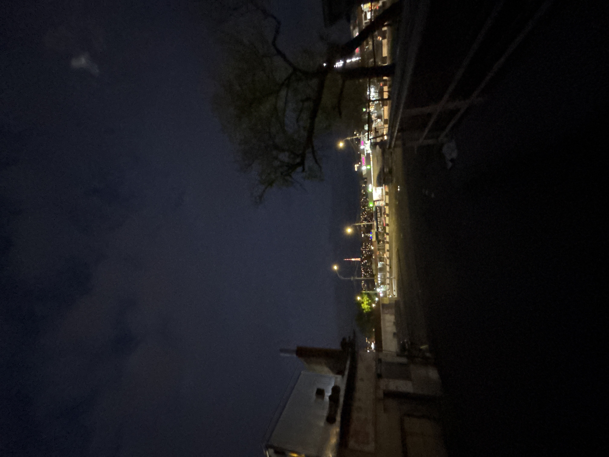 The image displays a nighttime urban scene in Yerevan, Armenia, captured with a landscape orientation. The dominant feature is the stark contrast between a dark, cloudy sky and a brightly illuminated street scene stretching across the midground.
The upper portion of the frame is dominated by a deep indigo-black sky, heavily overcast with visible cloud formations. A faint, irregular, and diffuse whitish glow is discernible in the upper left quadrant, possibly light reflecting off distant clouds or a celestial body obscured by haze.
The central and right-hand side of the image (when viewed horizontally) reveals a bustling city street or boulevard. A continuous line of artificial lights, including streetlights with bright, concentrated beams, and illuminated windows and signs from various buildings, creates a vibrant horizontal strip of light. These lights suggest the presence of shops, businesses, or residences lining the thoroughfare. Some lights appear blurred, hinting at movement or lens effects.
A large tree, with its dark, gnarled branches and sparse foliage, is silhouetted prominently against the illuminated cityscape and the slightly lighter sky areas. In the bottom-left corner, a partial view of a building's rooftop is visible, featuring some pipes or vents. The immediate foreground on the right is almost entirely obscured in deep shadow, appearing as an indistinct, dark mass, possibly a raised ground level or an adjacent structure.
No people or vehicles are distinctly visible, nor is any text discernible. The overall atmosphere is that of a quiet, late-night cityscape, defined by the glowing artificial lights against the dark, cloudy night sky.