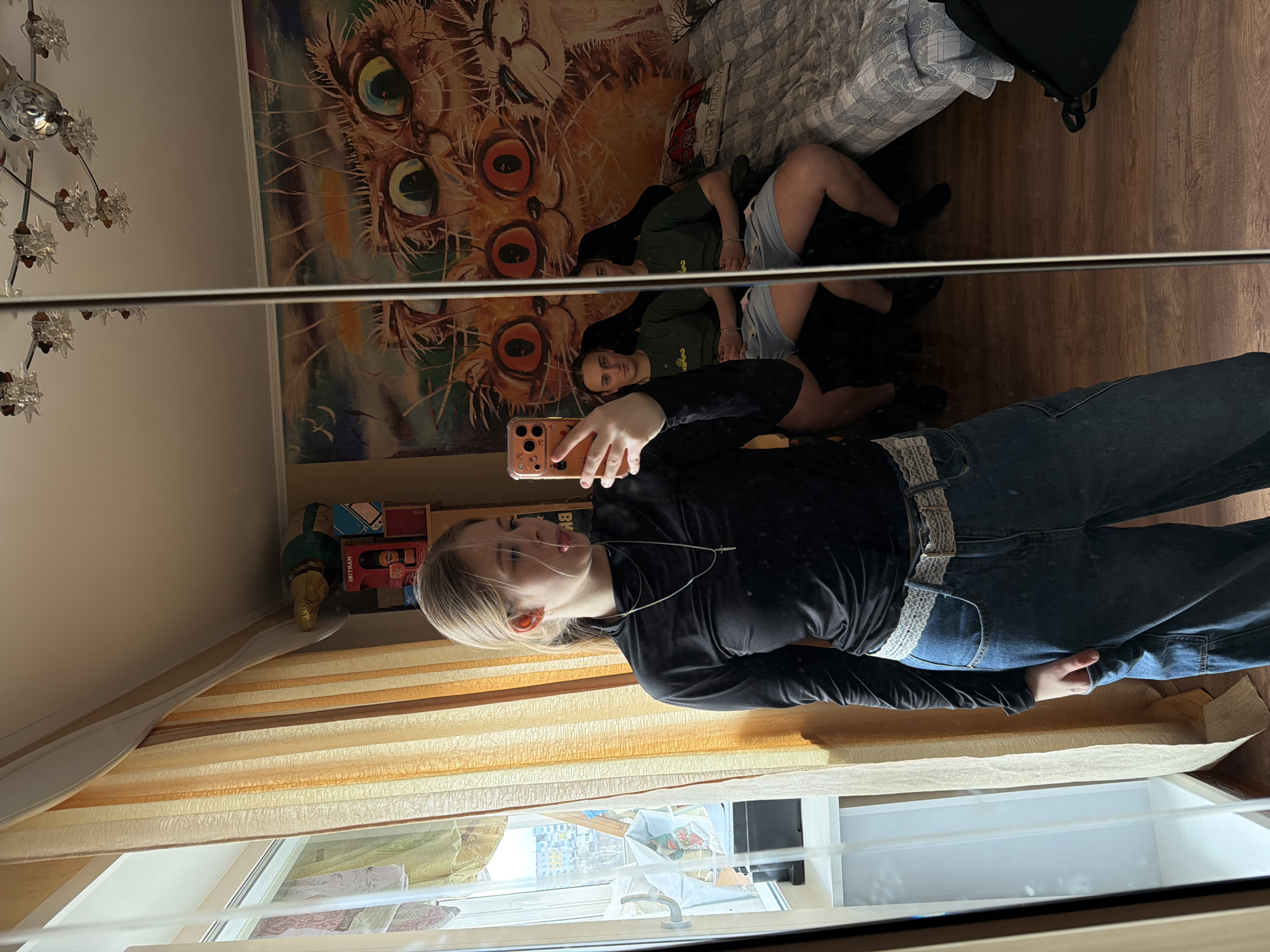 A mirrored selfie shows a young woman in a black long-sleeved shirt and jeans with a wide, decorative belt. She holds a phone to take the picture, reflecting two other people sitting on a bed under a comforter. A large, colorful cat-themed tapestry adorns the wall behind the bed. The reflection also shows a window with yellow and brown blinds, and a glimpse of the outside view suggests a cityscape. A chandelier is partially visible in the upper left corner. A shelf with various items sits to the left of the reflection of the woman. The photo appears to be taken indoors, possibly in an apartment or house in Perm, Russia, based on the architectural style and language seen outside the window.