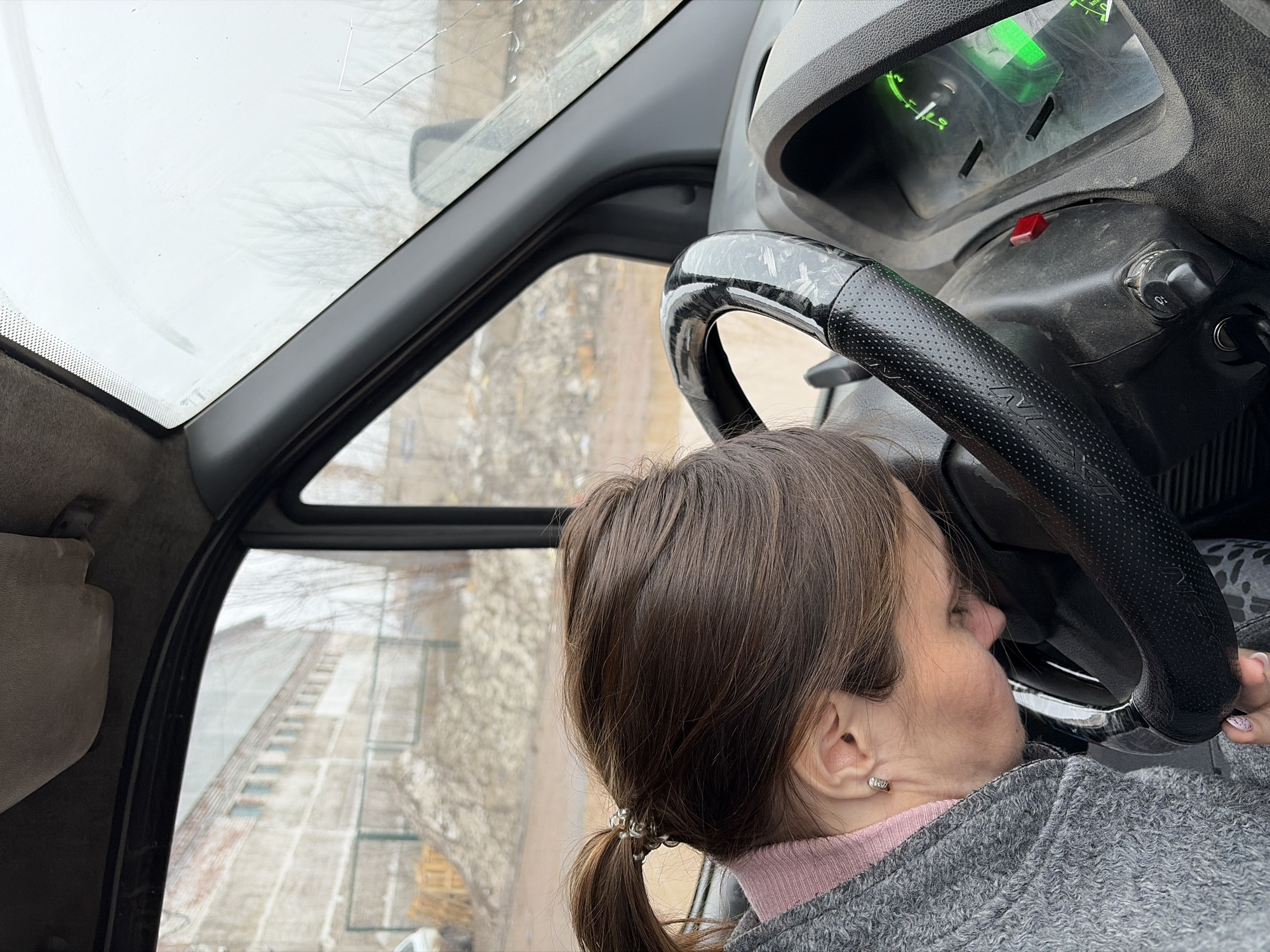 A woman with brown hair pulled back in a ponytail is seen inside the cab of a vehicle. Her head is leaning against the steering wheel. The vehicle's dashboard and window are visible. The outside scene through the window shows a city landscape that can be identified as Izhevsk, Russia.