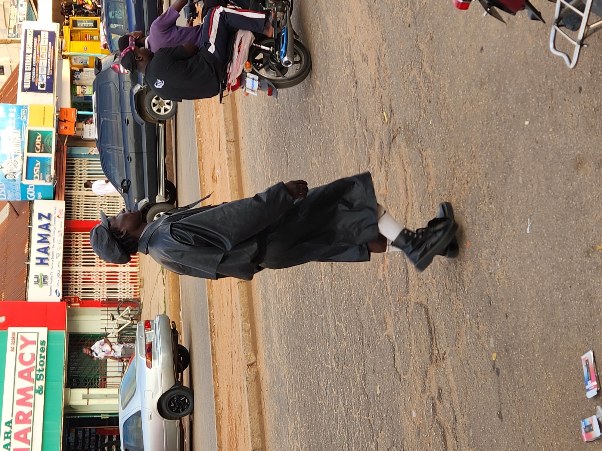 The image shows a street scene in what appears to be a commercial area. A man in a black coat and cap walks on the sidewalk next to the street. Buildings and shops line the street with signs visible, including "ARA PHARMACY" and "HAMAZ". A silver car is parked near the pharmacy, and other vehicles including a blue SUV and a motorcycle with riders are also present in the scene. The road is asphalt. The image seems to be taken at an angle, making the scene appear somewhat tilted. Discarded objects, possibly product packaging, are scattered on the ground.