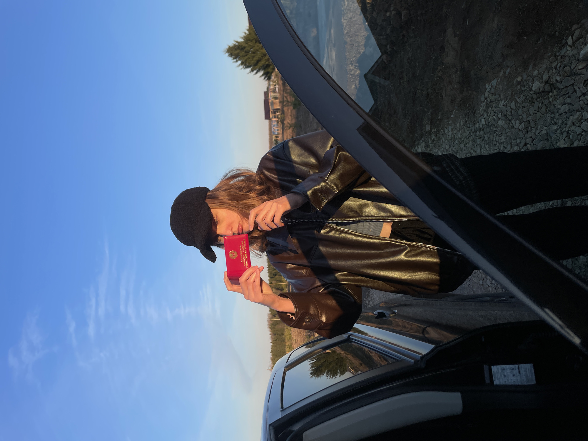 A young person with long brown hair, wearing a black newsboy cap and a dark brown faux leather jacket, stands next to a dark-colored car with its trunk or rear door open. They are holding a red document, identifiable as a passport, up to their face. Visible text on the document includes "РОССИЙСКАЯ ФЕДЕРАЦИЯ" (Russian Federation) and "ПАСПОРТ" (Passport) in Cyrillic.
Inside the open car compartment, several stacks of banknotes are visible. The setting is outdoors under a clear, bright blue sky with some faint cirrus clouds. The ground is rough, covered with gravel and dirt. In the background, there are trees and distant buildings, suggesting a rural or semi-rural landscape. The warm, soft lighting indicates either late afternoon or early morning. The overall scene suggests a travel stop or a moment during a journey in Podshivalovo, Russia.
