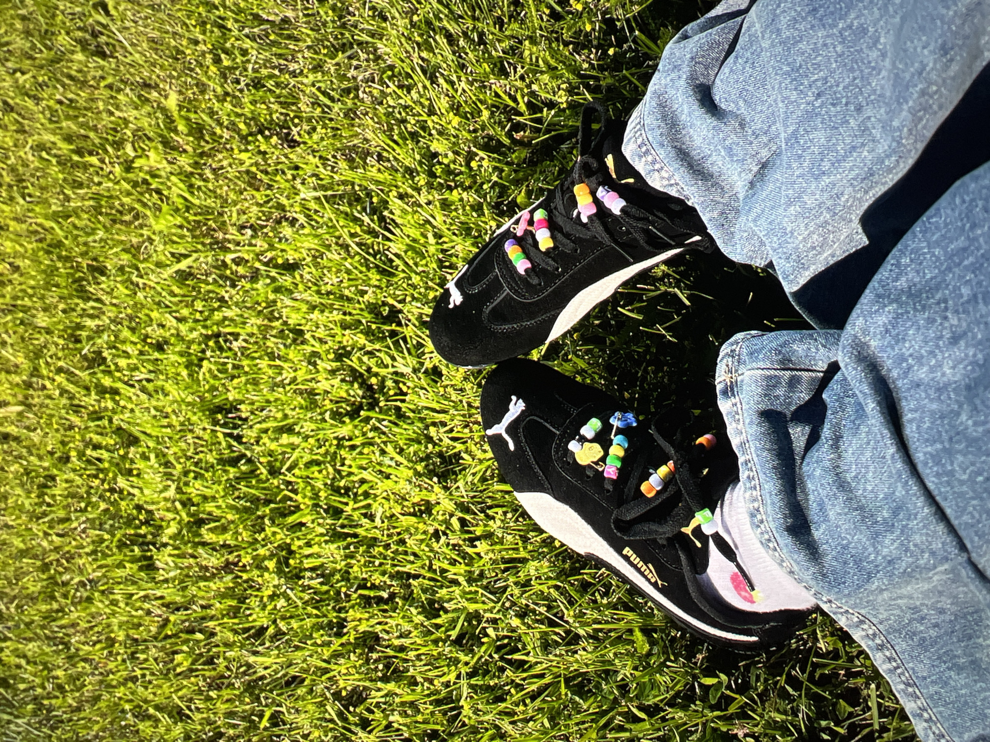 Two black Puma sneakers are on a bright green lawn. The sneakers have colorful beads threaded onto the laces. The person wearing the shoes is wearing blue jeans. The scene appears to be outdoors on a sunny day.