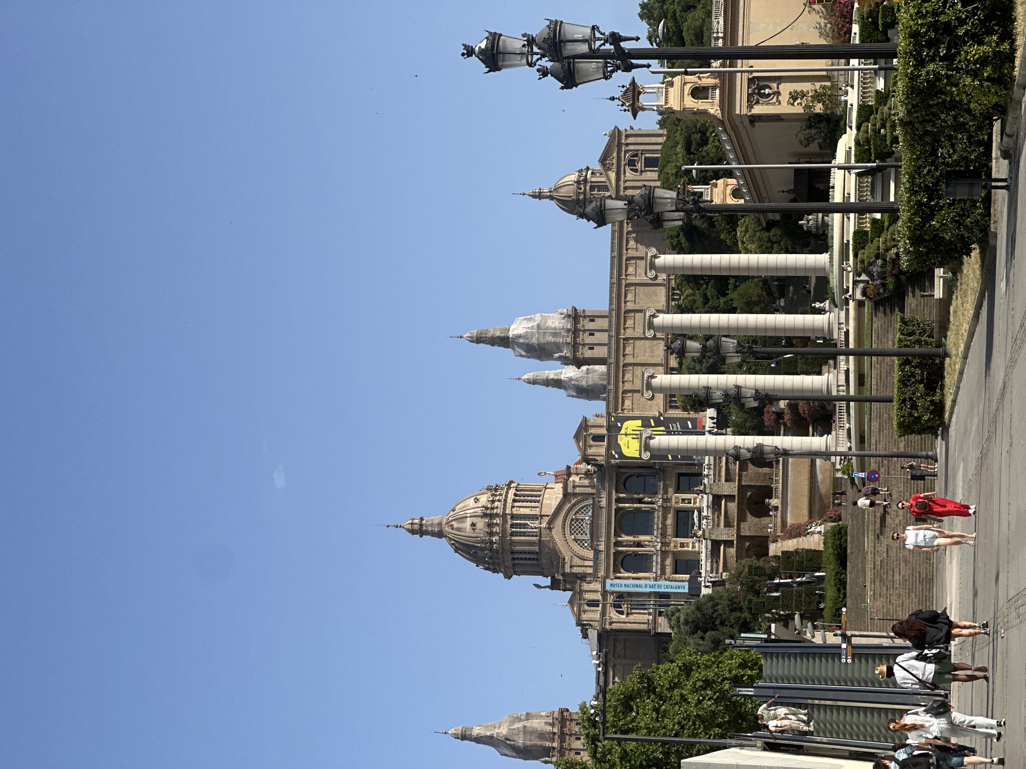This is a vertical, full-shot photograph taken on a bright, sunny day in Barcelona, Spain. The image showcases the grand architecture of the Palau Nacional, with its iconic domes and columns, set against a clear blue sky. A pathway and manicured greenery lead up to the building, and ornate lampposts line the walkway.
Several people are visible, walking along the pathway. One woman in a vibrant red dress stands out, while others are dressed casually. Some appear to be tourists, taking in the sights. The time of day is likely midday, given the strong sunlight casting shadows.
Visible text includes a blue sign that reads "MUSEU NACIONAL D'ART DE CATALUNYA," indicating the building's identity as the National Art Museum of Catalonia. The overall atmosphere is one of a pleasant, leisurely day in a historically rich and visually impressive urban setting.