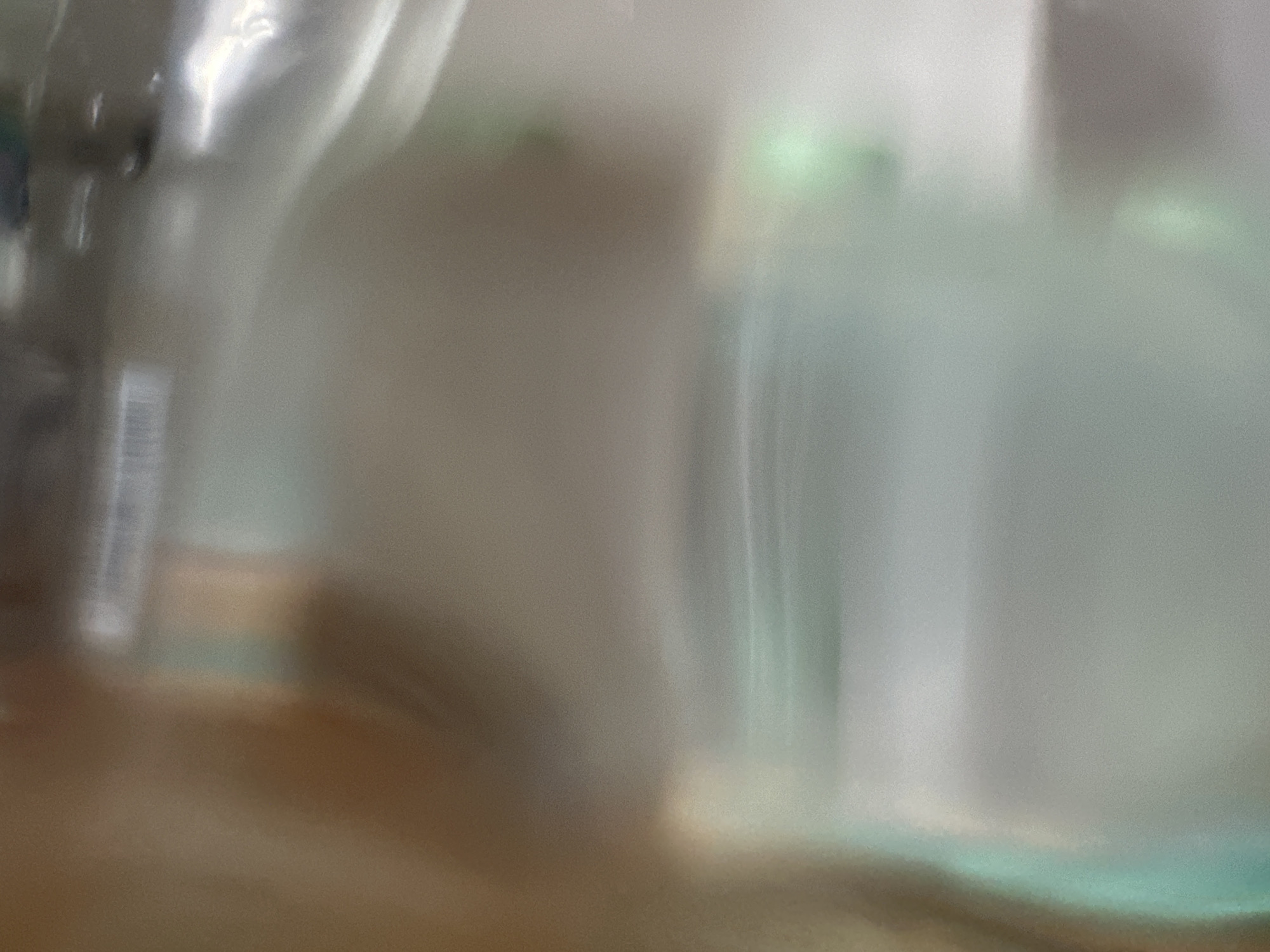 This media file captures a blurred, abstract scene, possibly through a frosted or textured glass surface. The primary subjects are indistinct, appearing as soft, pale shapes. On the left, a lighter, vertical object with some subtle horizontal lines is visible, perhaps a bottle or container. To its right, a larger, amorphous form dominates the center, lacking clear definition. Further to the right, there are suggestions of other objects or surfaces, with streaks of pale green and white light, hinting at reflections or internal illumination. The background is a muted blend of browns and grays, creating a soft, diffused atmosphere.
There is no clear activity or event depicted; the image seems to be a still life or a study of light and form rather than a narrative scene. Notable details are difficult to discern due to the blurriness, but the lighting suggests an indoor setting, possibly with artificial light sources contributing to the bright highlights and soft glows. The color palette is muted, with dominant grays, whites, and browns, punctuated by pale green accents. There are no discernible textual elements in the image. The overall impression is one of quietude and mystery, with the abstract nature of the image inviting interpretation. Given the location context of Belgrade, Serbia, this could be an interior shot of a home, a shop, or an office, viewed through a window or partition.