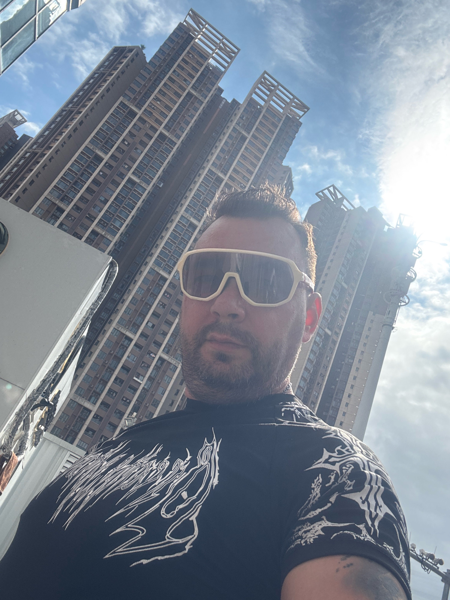 In a selfie taken outdoors in Nanhai District, China, a man with a beard and sunglasses faces the camera, his head and shoulders dominating the foreground. He is wearing a black t-shirt with a white graphic print, and his right arm, partially visible, shows tattoos. The background is dominated by a bright sky with some clouds and the imposing architecture of tall, modern buildings. The buildings are angled and appear to be under construction or recently built, with visible scaffolding and repetitive window patterns. The sun is high in the sky, creating a strong glare in the upper right portion of the image.