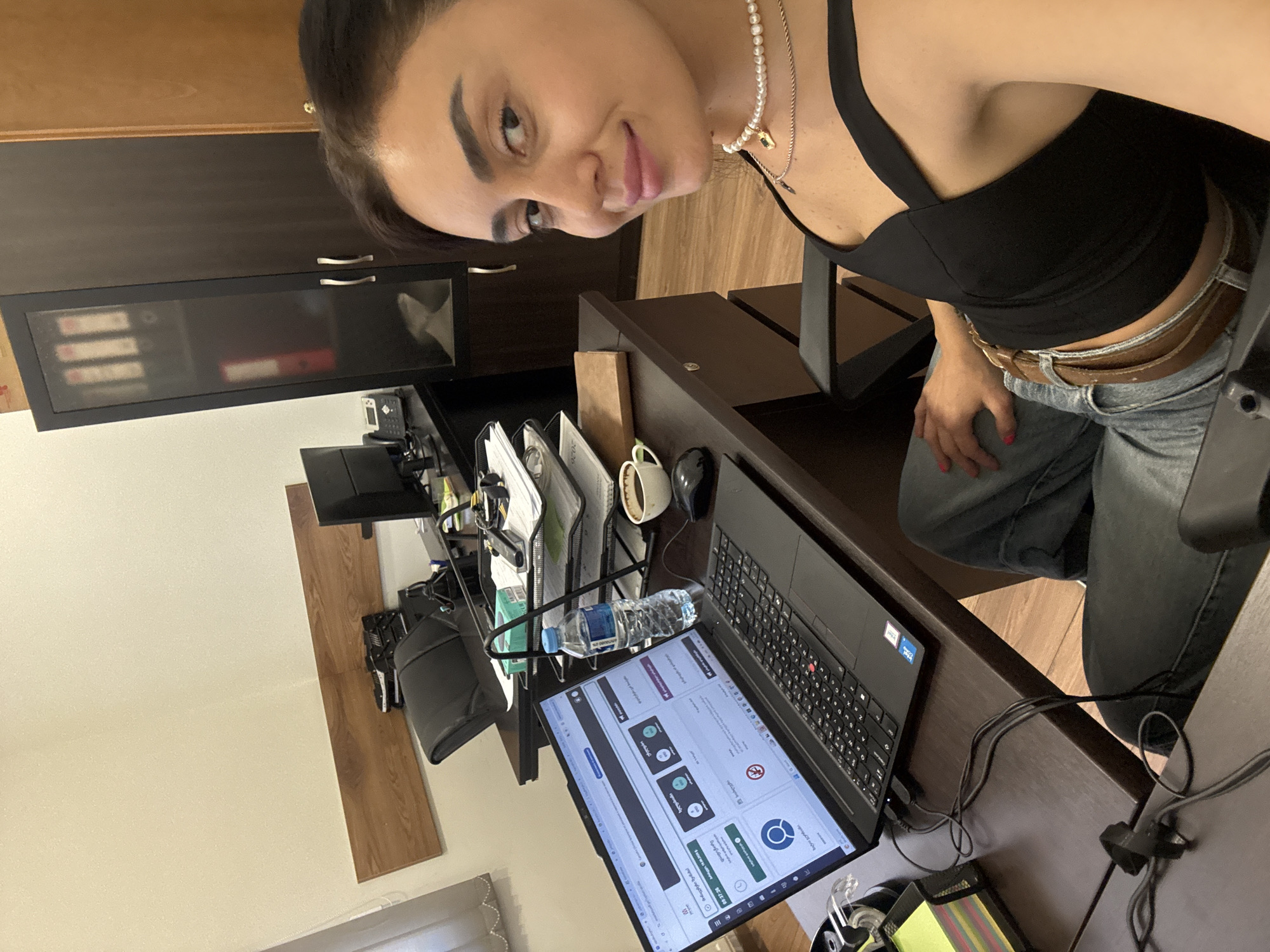 The image depicts a woman sitting at a desk in what appears to be an office environment in Tbilisi, Georgia. She is looking towards the camera and smiling. On the desk in front of her is an open laptop displaying a website with Georgian text, a coffee cup, a bottle of water, and a computer mouse. Behind the desk, a filing cabinet with glass doors containing binders is visible. The woman is wearing a black top and jeans. The overall scene suggests a work setting within a city.