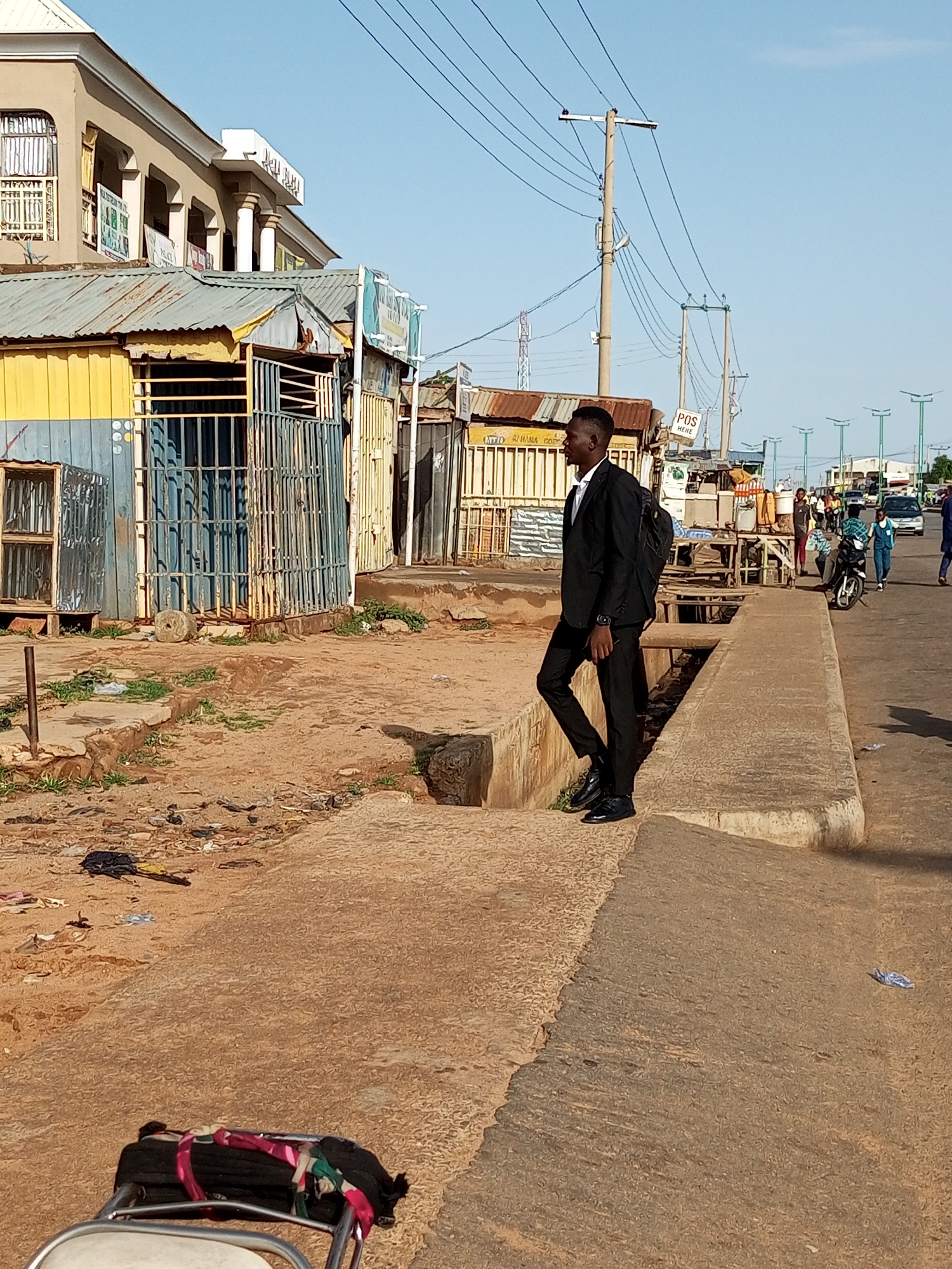In Garko, Nigeria, a young man in a black suit and backpack stands on a concrete ledge beside a dirt path. The path is littered with trash, and a metal railing lies in the foreground. Behind the man, corrugated metal structures with barred windows line the street. In the distance, the street is busy with pedestrians, motorbikes, and cars, all under a clear blue sky. Electrical poles and wires are visible, with streetlights in the far distance. The overall scene depicts a bustling urban environment with elements of informal commerce and daily life.