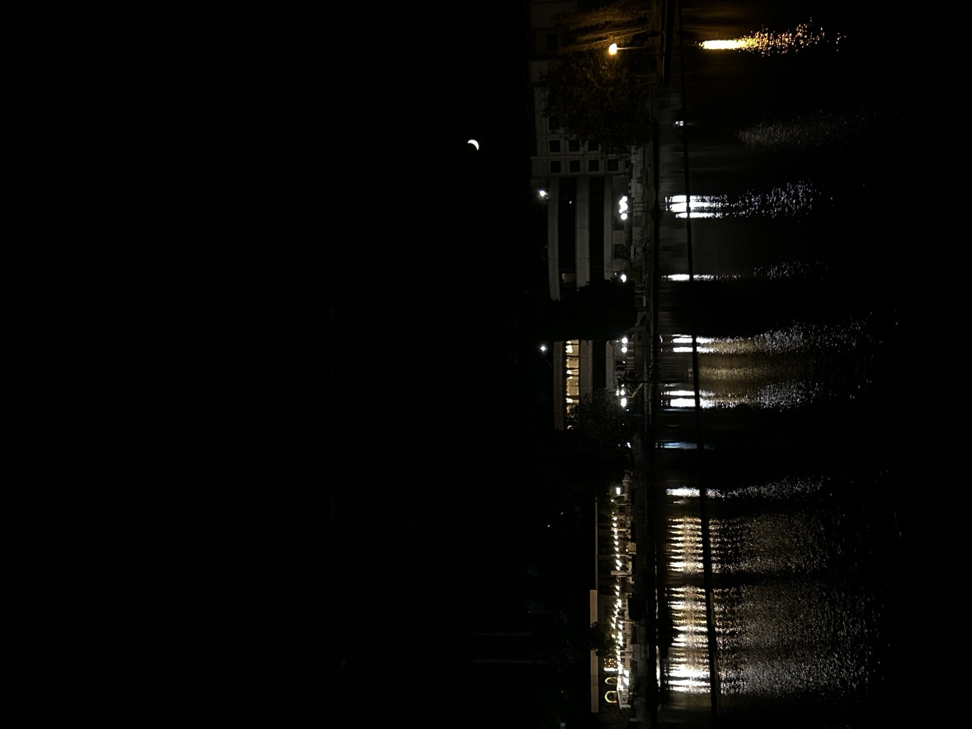 The image depicts a night scene in an urban environment, possibly near a body of water. A crescent moon is visible in the sky. Along the right side of the frame, there are building structures with illuminated windows and streetlights. The reflections of these lights shimmer on the water's surface. The left side of the image is predominantly dark. The location appears to be a city at night, but the specific city is unknown.