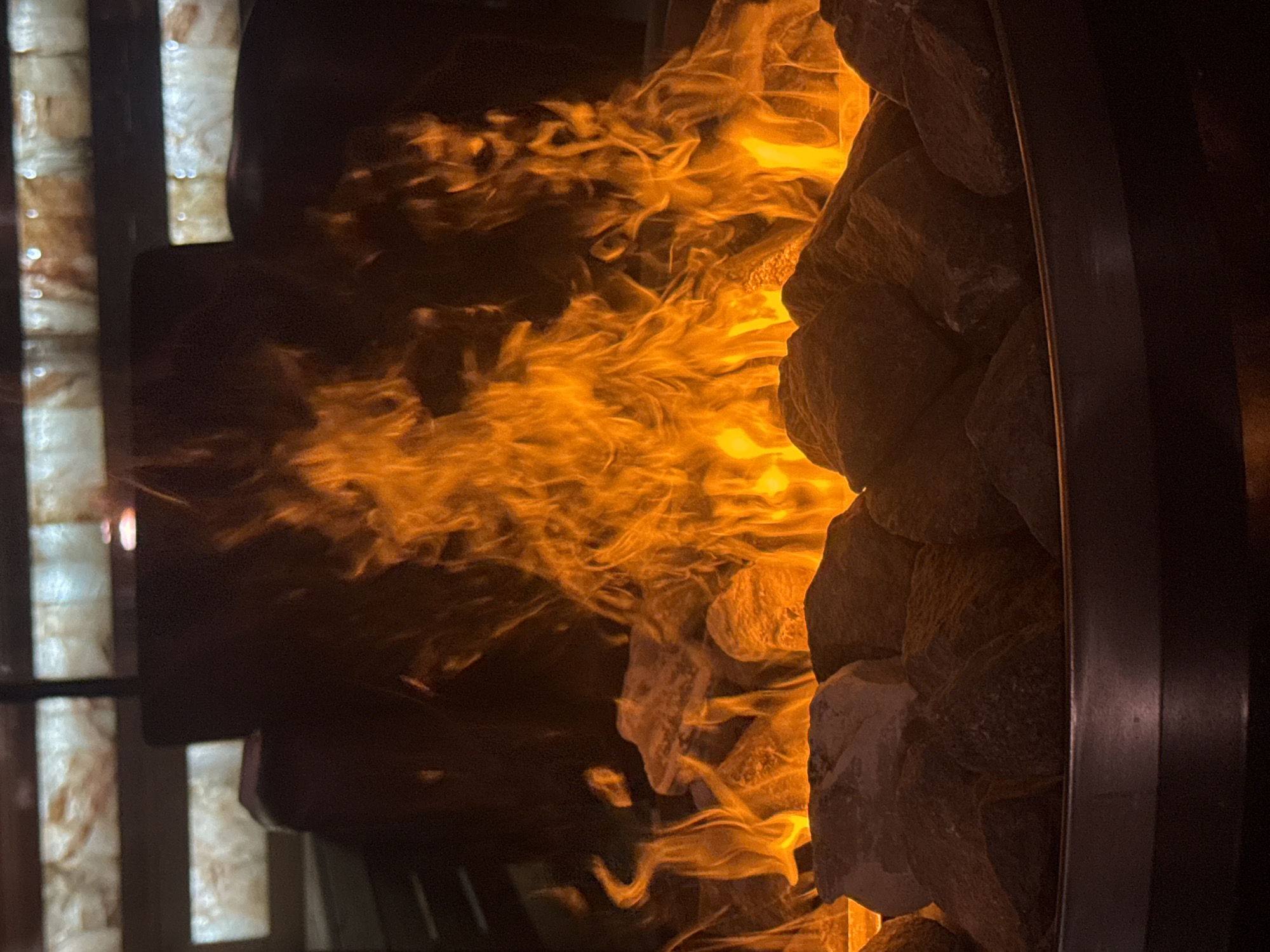 The image depicts a close-up view of a fire feature in Plovdiv, Bulgaria. The central focus is a vibrant, multi-hued orange and yellow flame rising dynamically from a bed of dark, irregularly shaped rocks. These rocks are contained within a dark, metallic, circular basin, the upper rim of which is visible in the lower foreground. The flames illuminate the surrounding rocks, casting a warm glow. In the background, above the fire, multiple rectangular panels, appearing to be translucent stone such as onyx with visible veining, are internally illuminated, emitting a soft, diffused light. These panels are arranged horizontally, forming a decorative architectural element. Between the fire and the illuminated panels, indistinct dark shapes are visible, appearing as blurred architectural or furniture elements. No individuals are present within the frame.