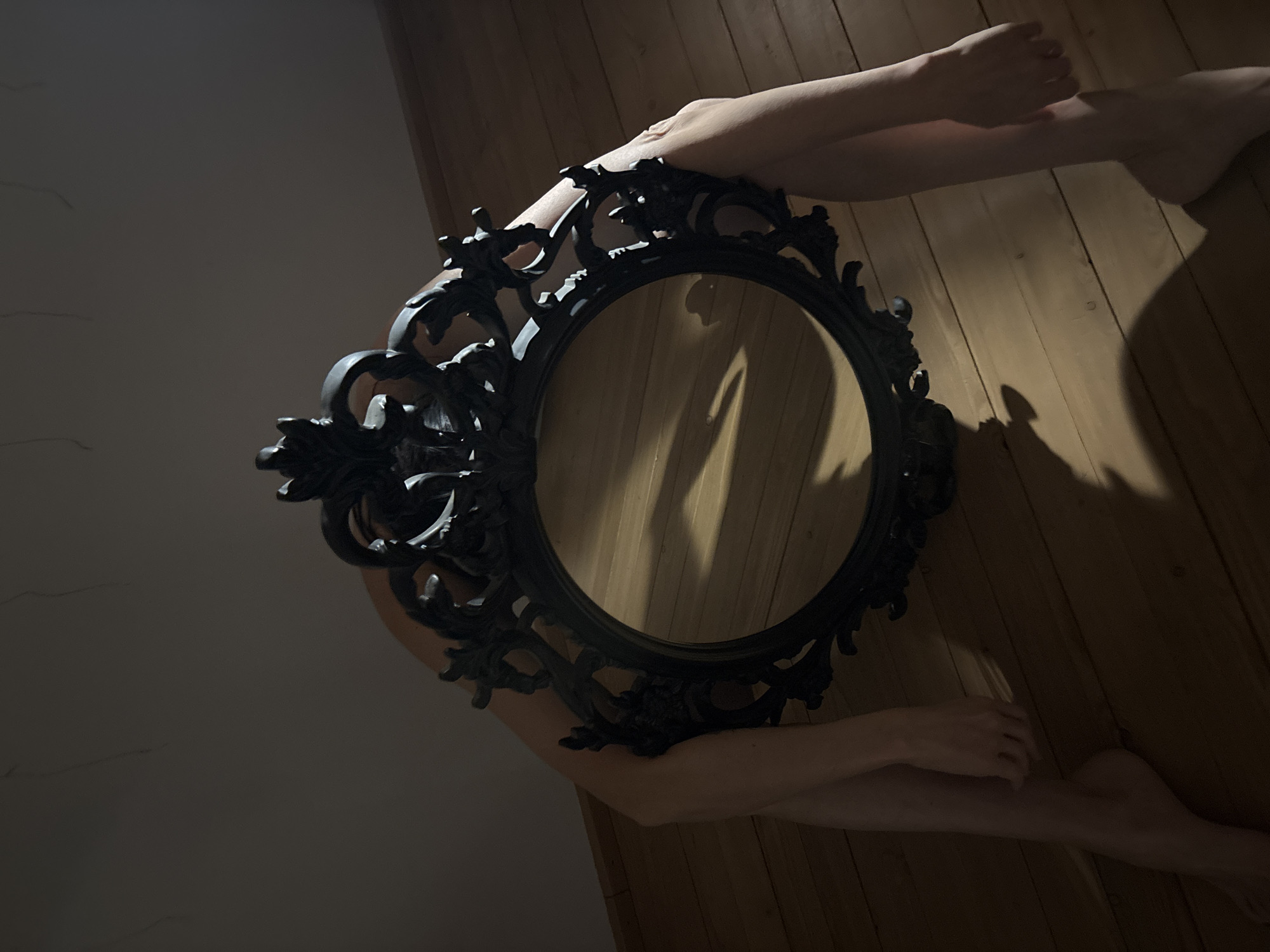 A low-angle shot depicts a person obscured behind a black, ornate, circular mirror lying on a wooden floor. The person's arms and legs extend from behind the mirror's edges. The scene is partially lit, casting shadows from the mirror and the person's limbs onto the floor and the adjacent wall. The environment appears to be indoors, possibly a room with bare walls and wooden flooring. The image's location in Chaikovsky, Russia, is unconfirmed by the visual data.