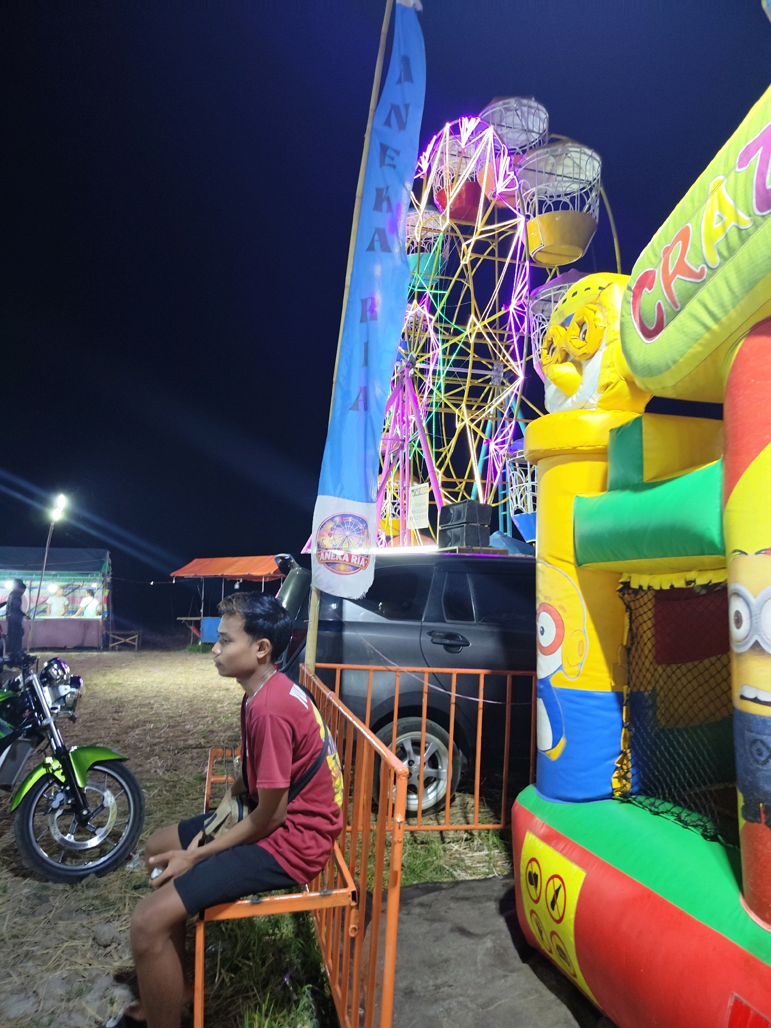 The image depicts a nighttime scene at an outdoor amusement area in Suling Wetan, Indonesia. A young man, appearing to be in his late teens or early twenties, sits on an orange metal bench in the foreground, facing to the left. He is wearing a red t-shirt and black shorts, with a tan bag slung across his chest. To his left, a green motorcycle is parked on a grassy, uneven ground.
In the mid-ground, a dark-colored SUV is positioned behind a row of orange fencing. Behind the SUV, a Ferris wheel illuminated with colorful lights is prominently displayed against the night sky. A large, inflatable structure, primarily yellow, green, and red, with depictions of cartoon characters, is situated to the right of the frame. A blue flag with writing and a circular emblem is also visible, partially obscuring the Ferris wheel. In the background, tents and stalls are set up, with lights indicating a public gathering or market. The overall environment suggests a festive atmosphere at a local fair or carnival.