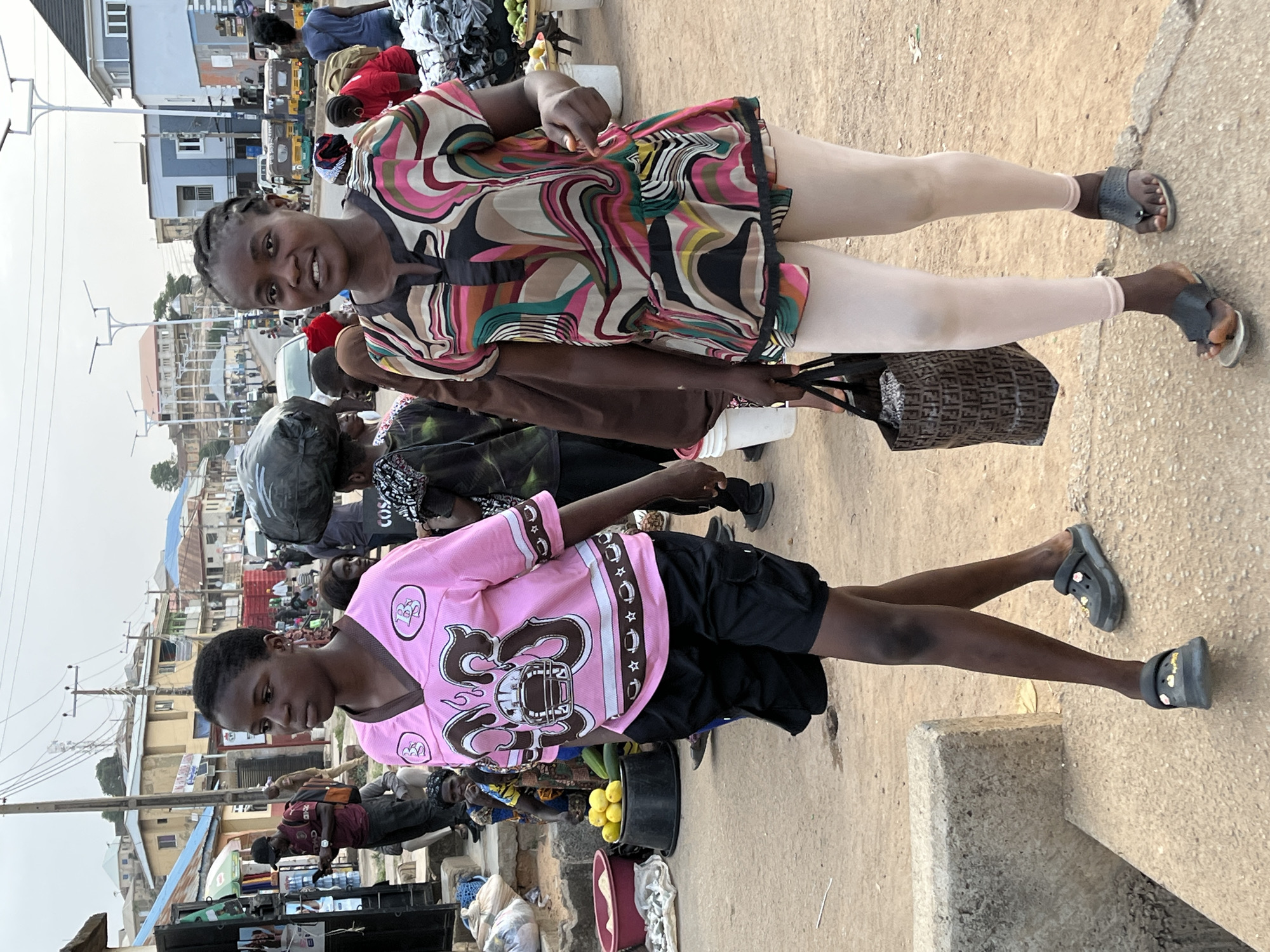 Two young adults stand in an outdoor market setting in Jos, Nigeria. The market is bustling with people and goods.
The first individual, a young woman, faces the camera, smiling. She has braided hair and wears a multi-colored, patterned, short dress with skin-toned leggings and dark sandals. She carries a dark-colored woven bag.
The second individual, a young man, looks downward, away from the camera. He has short hair and wears a pink shirt with an American football-themed graphic on the front, paired with black shorts and black Crocs-style shoes. A bucket of fruit (likely lemons or oranges) sits near him.
The environment includes market stalls and various vendors. There are buildings and power lines visible in the background. The ground is dusty and uneven. People are carrying items and going about their business.