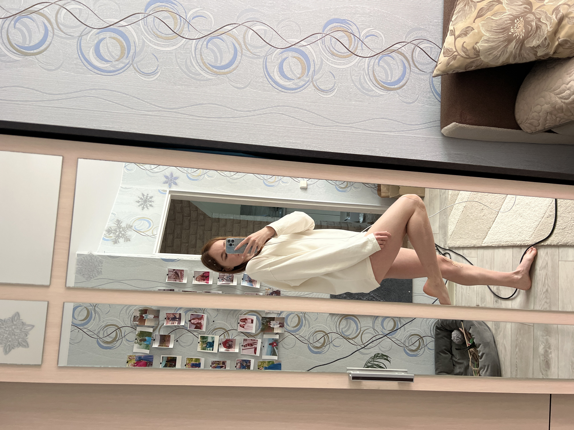 A woman is reflected in a mirror. She holds a phone in her left hand, facing the screen toward herself, taking a photo. She is wearing a long-sleeved, cream-colored shirt. Her right leg is extended, and her left leg is bent at the knee. The background behind her includes a door frame and a wall with decorative snowflakes and horizontal lines. Below her reflection are small, printed photographs attached to the wall above the mirror. The room is indoors, with walls covered in patterned wallpaper with circles and wavy lines. A section of a couch with pillows is visible at the top right of the frame. Black wiring is also visible. It is not possible to determine the location is in Chaikovsky, Russia.