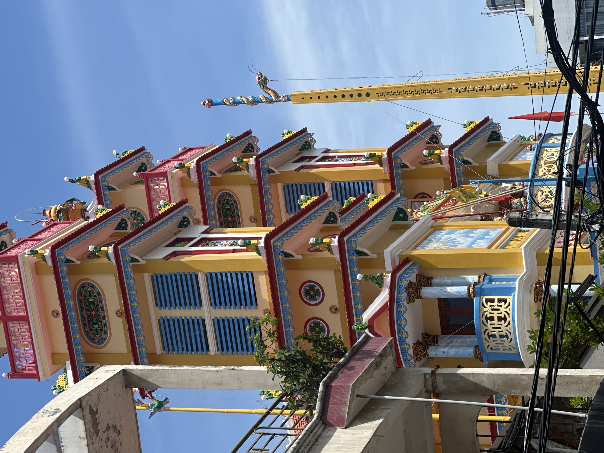 The image shows a brightly colored, multi-tiered pagoda structure, painted in vibrant yellow, red, and blue. The pagoda is adorned with intricate decorative elements, including patterned friezes, circular motifs with floral designs, and small statues of mythical creatures. Several levels of the pagoda feature balconies with ornate railings and decorative pillars. To the right of the pagoda, a tall, yellow pole stands, topped with a blue and red ornamental spire and a dragon sculpture. The structure is situated in an urban environment, with a partial view of an adjacent building made of concrete and metal railings to the left. Electrical wires are visible in the foreground, spanning across the lower part of the image. A red flag is also visible near the base of the yellow pole. The sky is clear and blue. The pagoda is identified as being located in Nha Trang Ward, Vietnam.