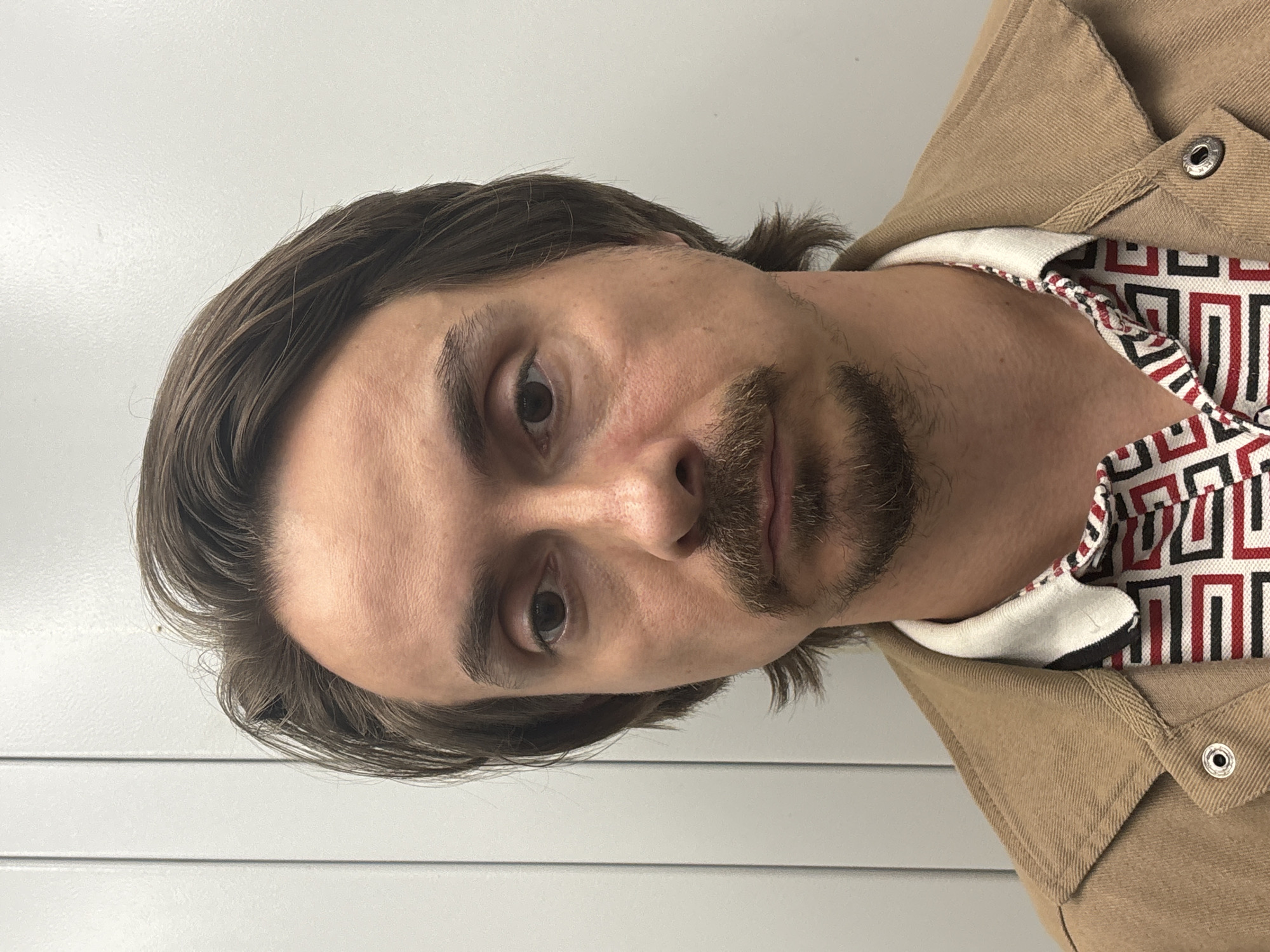 A close-up, chest-up, medium shot depicts a Caucasian male facing forward against a plain, light-colored, vertically grooved wall. The man has medium-length, brown hair styled back from his forehead, with some wisps falling forward. He has a light brown mustache and a goatee. His skin appears fair with some redness around the cheeks and under his eyes. He is wearing a tan jacket with a visible snap closure on the left side of the frame. Underneath the jacket, he wears a collared shirt with a geometric pattern of black, white, and red shapes. The collar of the shirt is white with black and red trim. The lighting is even and bright, with no harsh shadows. The overall impression is that of a mugshot or official photograph.