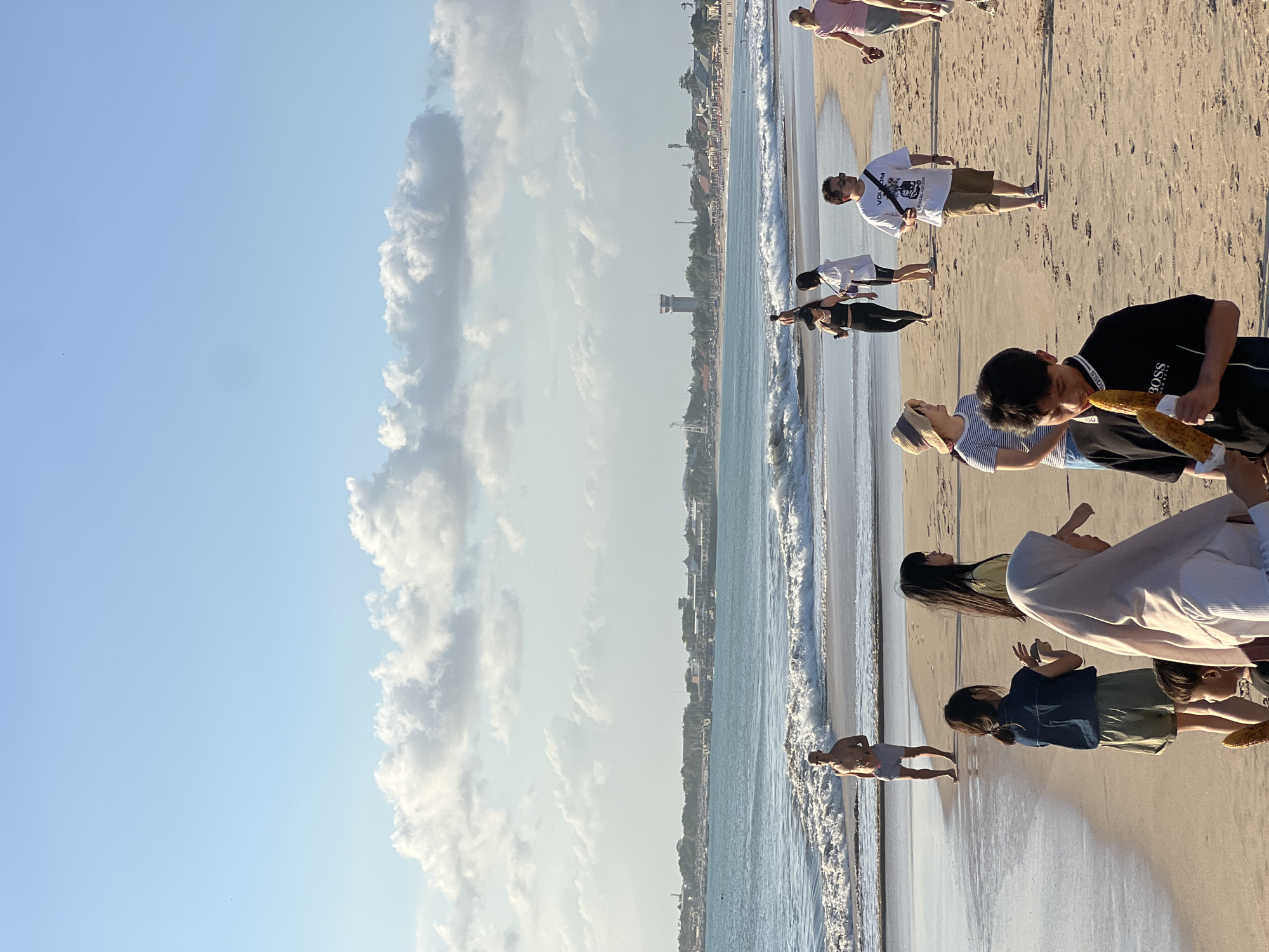 The image depicts a beach scene in Kuta Selatan, Indonesia. The foreground features several people on the sandy shore. In the center, a young boy in a black BOSS t-shirt is holding and looking at two ears of corn. To his left, a young girl in a blue denim top and green shorts is standing, while a woman in a light-colored hijab is partially visible in front of her. Further to the left, a man in grey shorts is standing in the shallow water, facing the ocean.
In the middle ground, more people are walking along the beach. A group of four individuals, two women and two men, are walking together. One man wears a white t-shirt with graphics and brown shorts, another man is partially obscured, and two women are dressed casually. A woman in a straw hat and striped shirt stands near them.
The background shows the ocean with moderate waves, and beyond that, a coastline with buildings, palm trees, and a distinctive tower structure. The sky is a clear blue with scattered white, fluffy clouds. The lighting suggests it is late afternoon.
