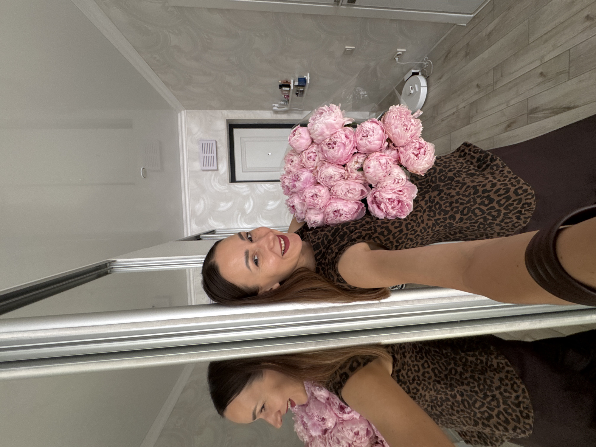 A woman is taking a selfie in a hallway, holding a large bouquet of pink peonies. She is smiling and looking at her reflection in a mirrored wardrobe. She is wearing a brown leopard-print top and a dark skirt. The hallway has textured wallpaper and wooden flooring. A white robot vacuum cleaner is visible on the floor in the background, near a wall outlet. A small shelf with toiletries is mounted on the wall. The image was taken indoors, likely in a residential setting.