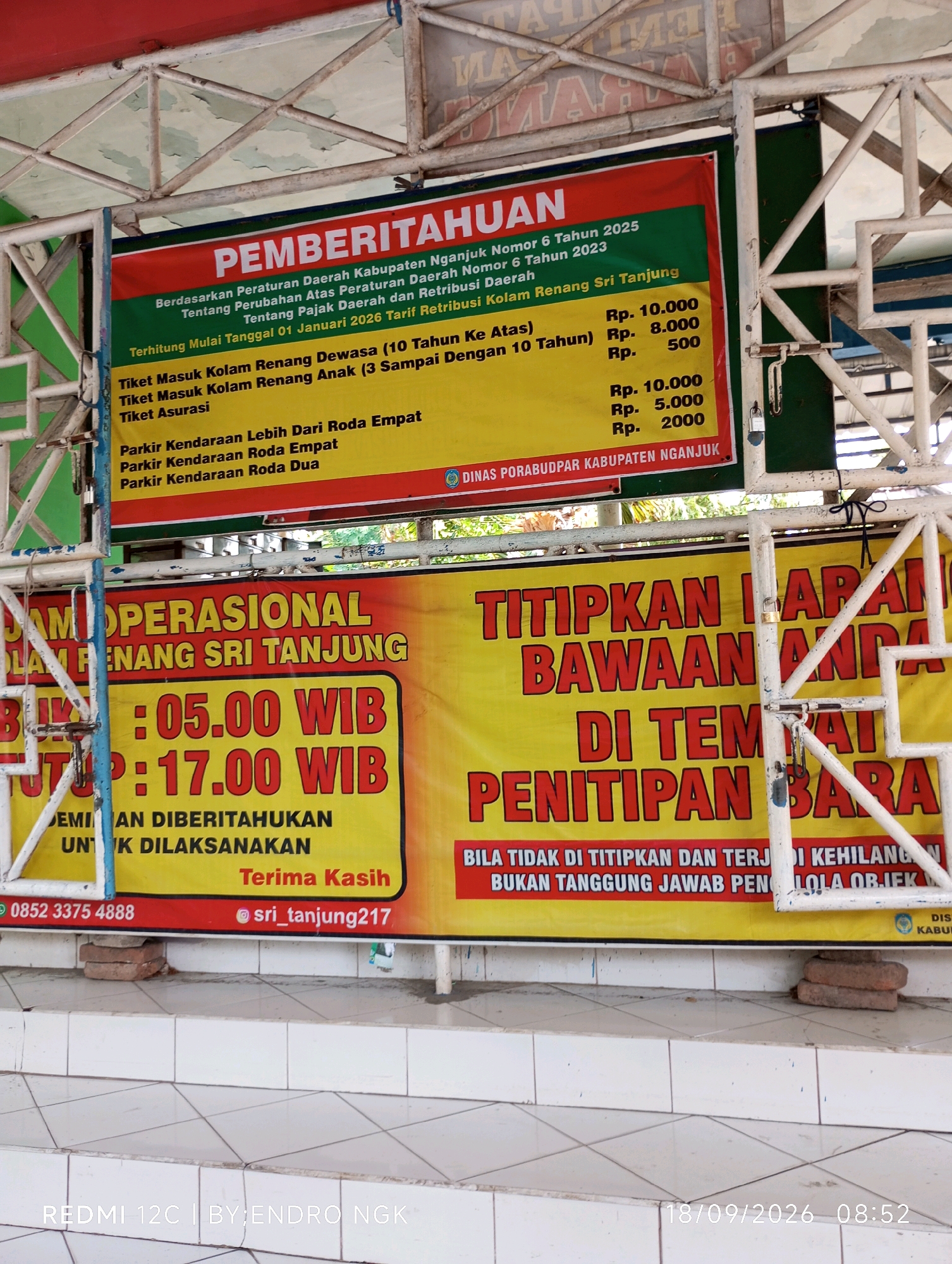 The image shows a public announcement board for the "Kolam Renang Sri Tanjung" (Sri Tanjung Swimming Pool) located in the city of Nganjuk, Indonesia. The board displays various information, including regulations, entrance fees, and operational hours. The main text at the top reads "PEMBERITAHUAN" (Announcement). Below this, it outlines regulations based on local government decrees regarding tax and retribution adjustments. Specific pricing is listed for "Tiket Masuk Kolam Renang Dewasa (10 Tahun Ke Atas)" (Adult Swimming Pool Entrance Ticket - 10 Years and Above) at Rp. 10,000, "Tiket Masuk Kolam Renang Anak (3 Sampai Dengan 10 Tahun)" (Child Swimming Pool Entrance Ticket - 3 to 10 Years) at Rp. 8,000, and "Tiket Asurasi" (Insurance Ticket) at Rp. 500. Parking fees are also itemized: "Parkir Kendaraan Lebih Dari Roda Empat" (Parking for Vehicles with More Than Four Wheels) at Rp. 10,000, "Parkir Kendaraan Roda Empat" (Four-Wheeled Vehicle Parking) at Rp. 5,000, and "Parkir Kendaraan Roda Dua" (Two-Wheeled Vehicle Parking) at Rp. 2,000. The announcement is attributed to "DINAS PORA BUDPAR KABUPATEN NGANJUK" (Nganjuk Regency Youth, Sports, Tourism and Culture Office). A second, larger banner, predominantly yellow with red lettering, announces "JAM OPERASIONAL KOLAM RENANG SRI TANJUNG" (Operational Hours of Sri Tanjung Swimming Pool). It states the pool is open from "05:00 WIB" (05:00 Western Indonesian Time) to "17:00 WIB" (17:00 Western Indonesian Time). It also includes a notice: "JEMINAN DIBERITAHUKAN UNTUK DILAKSANAKAN" (Guaranteed to be announced for implementation) and "Terima Kasih" (Thank You). Further down, another banner in red and yellow lettering states "TITIPKAN BARANG BAWAAN ANDA DI TEMPAT PENITIPAN BARANG" (Deposit Your Belongings in the Luggage Deposit Area). It includes a disclaimer: "BILA TIDAK DI TITIPKAN DAN TERJADI KEHILANGAN BUKAN TANGGUNG JAWAB PENGELOLA OBJEK" (If not deposited and a loss occurs, it is not the responsibility of the object manager). Contact information is visible at the bottom left of the announcement board: a phone number "0852 3375 4888" and an Instagram handle "@sri_tanjung217". The overall scene appears to be outdoors, with a metal structure supporting the banners. The environment suggests a public facility, possibly an entrance area to the swimming pool. The lighting indicates daytime. There are no people clearly visible in the image, only the signage. The image is a photograph taken from a slightly low angle, capturing the full extent of the banners and the supporting structure. The lower portion of the image shows tiled stairs and parts