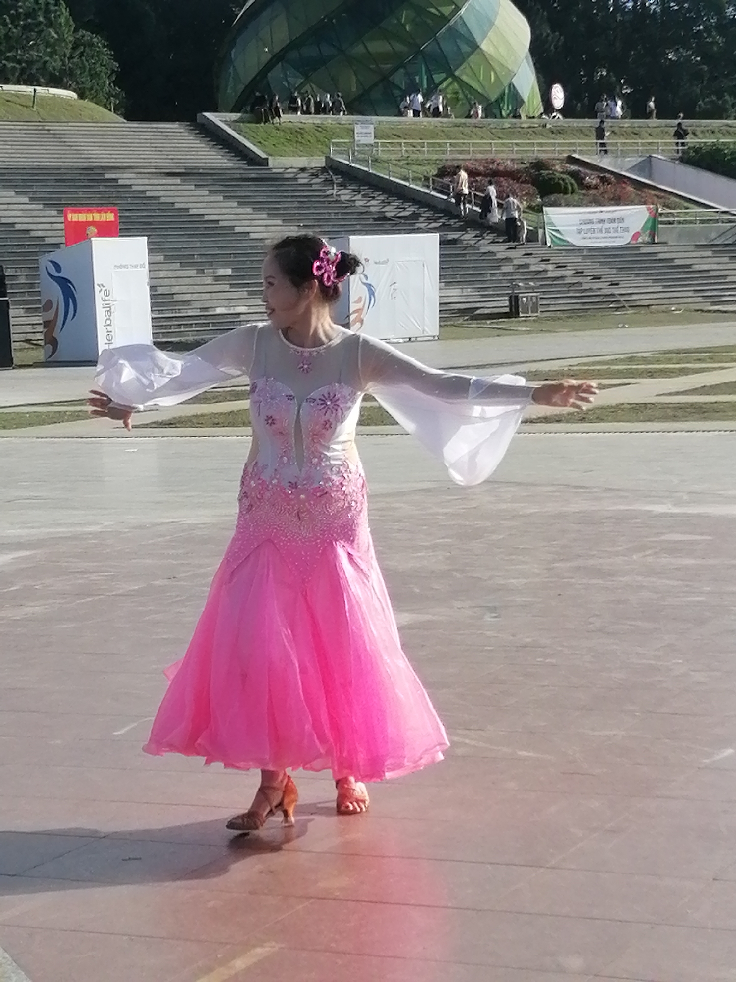 A woman in a pink ballroom dancing dress with sheer white sleeves is performing a dance outdoors. The dress is fitted at the bodice, adorned with intricate beadwork and floral embellishments, and flares out into a voluminous skirt. Her hair is styled up with a pink flower accessory. She is wearing tan heels.
The setting appears to be an open paved area, possibly a plaza or dance floor, with a large, stepped amphitheater in the background. Behind the amphitheater, there are trees and a distinctive geodesic dome structure with green and yellow panels. Several people are visible in the background, some seated on the steps of the amphitheater and others walking around. The lighting suggests it is daytime, with shadows indicating sunlight.
One of the objects in the background is a white rectangular structure with a blue logo and text that appears to read "Herbalife". A red banner is also visible on top of this structure. Another banner further back displays Vietnamese text. The overall atmosphere seems to be one of performance or a public event, with a sense of dynamism from the dancer's pose. The location is identified as Da Lat, Vietnam.