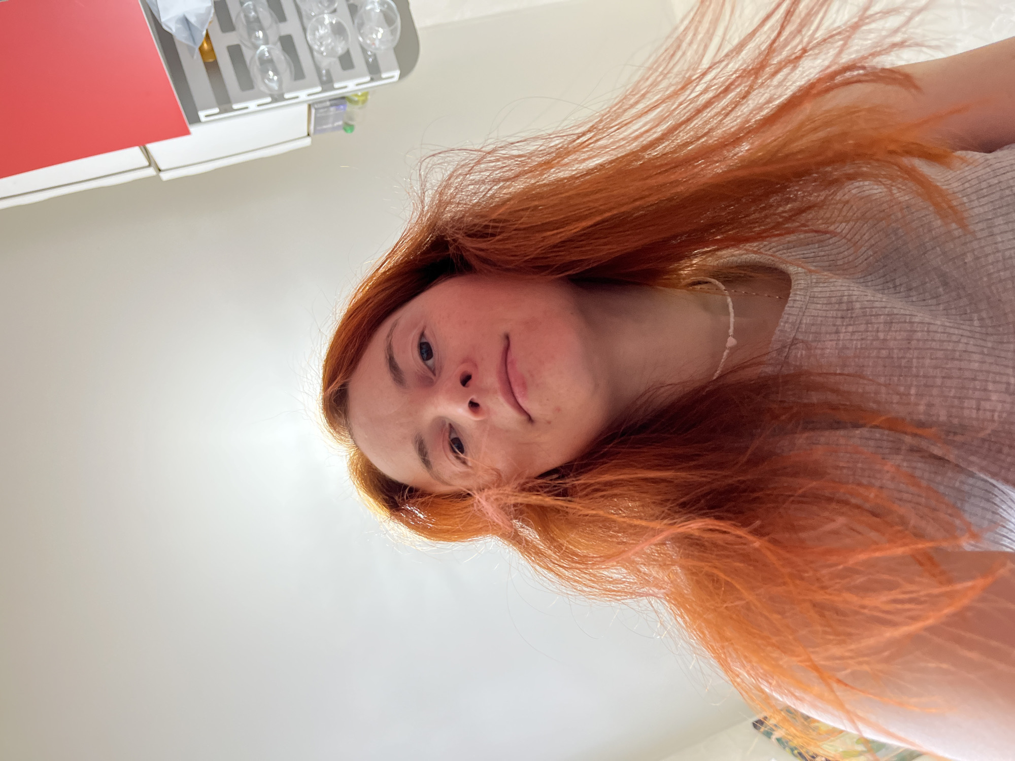 A low-angle, slightly rotated shot shows a fair-skinned woman with reddish-orange, long hair. Her head is tilted, with her face visible in the upper center of the frame. She has light-colored eyes, a small nose, and a neutral expression. A thin necklace is visible at her neck. She wears a light gray, horizontally ribbed shirt. The background is mostly a plain, off-white surface, possibly a ceiling or wall. In the upper left corner, a section of a white cabinet with a red door is visible, along with what appear to be several glass bottles or glasses in a rack. The location is unspecified, but the query suggests the city of Chaikovsky, Russia. The exact environment, actions, and interactions are difficult to discern given the limited context and framing of the image.