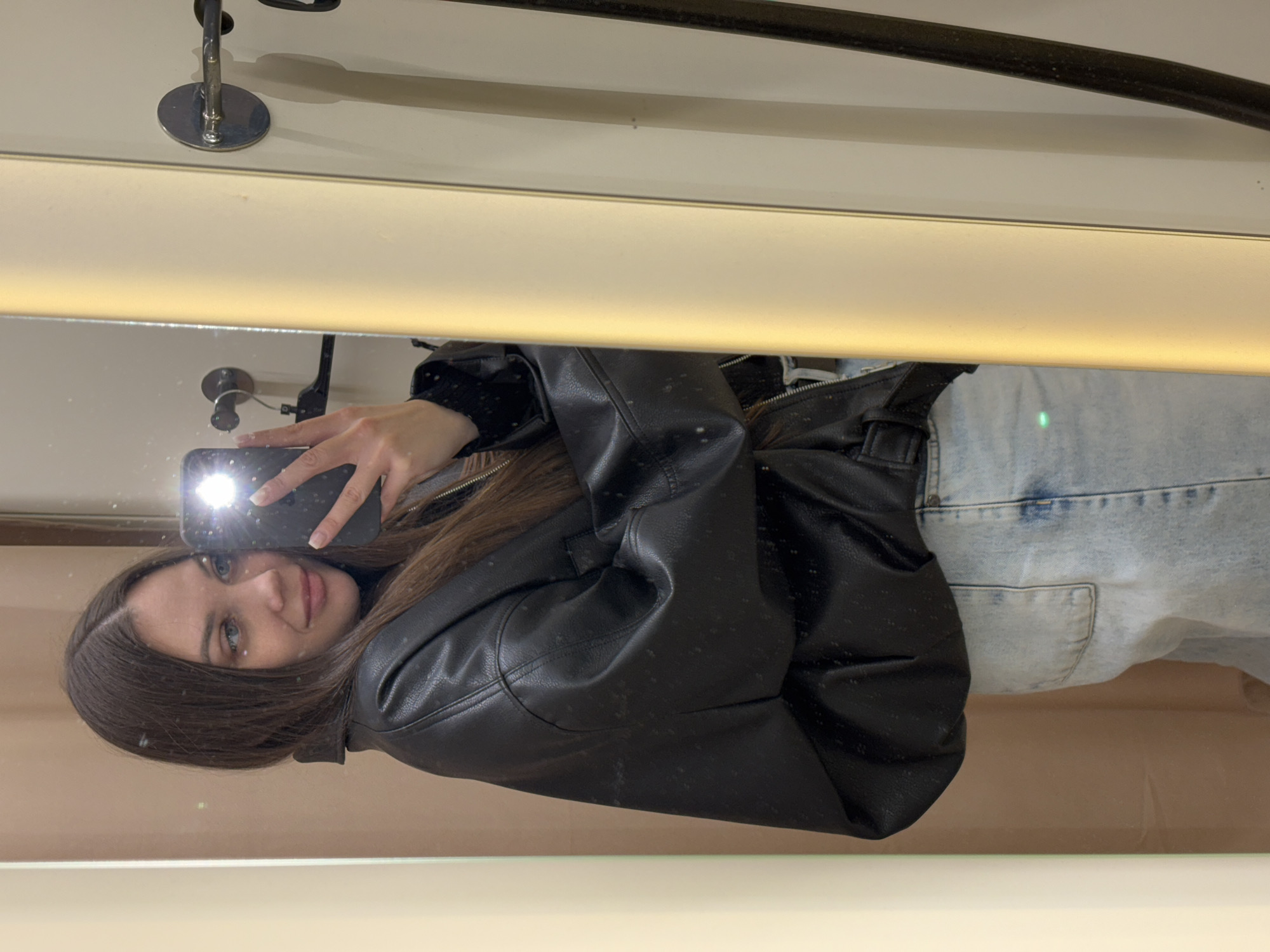 A mirror selfie captures a young woman from the chest up, reflected in a clean, vertical mirror. The image is rotated 90 degrees clockwise, showing the woman's head tilted to the right. She has long, dark brown hair parted down the middle and light-colored eyes. Her expression is soft and pleasant, with a slight smile.
She is wearing a black leather jacket over what appears to be a black top and light-wash denim jeans. Her right hand, with light-colored nails, holds a dark-colored smartphone, which is the source of a bright flash illuminating her face and creating a prominent glare on the phone's screen in the reflection.
The background shows the interior of a room, likely a fitting room or retail space. Above the woman's reflection, there's a horizontal light strip providing ambient illumination. Metal fixtures, possibly hooks or part of a clothing rack, are visible in the upper part of the mirror's reflection. The overall setting is indoors. No discernible text is visible in the image. The time of day is not indicated, and the location context is Yekaterinburg, Russia.