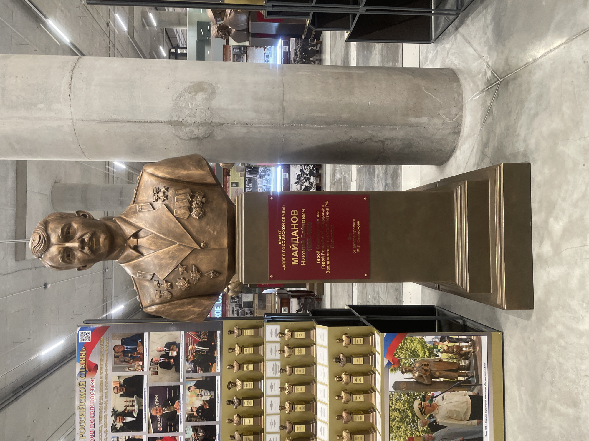 The image depicts a museum exhibit dedicated to Russian military figures. In the foreground, a bronze bust of a man in a military uniform stands on a pedestal. The bust is adorned with medals. Behind the bust, a large poster displays a collage of photographs of individuals in formal attire, some holding awards. To the left of the bust, a display case holds numerous smaller bronze busts on individual pedestals, each with a plaque. A large, concrete column is visible to the right of the bust. In the background, various exhibits and displays are visible, including what appear to be historical photographs and more busts. A Russian flag is also visible in the lower left corner of the image. The environment appears to be an indoor exhibition space with concrete floors and ceilings, illuminated by overhead lighting. The text on the red plaque in front of the bust reads "ПРОЕКТ «АЛЛЕЯ РОССИЙСКОЙ СЛАВЫ» МАЙДАНОВ Николай Саинович 1934-2000 Герой Советского Союза, Герой Российской Федерации, Заслуженный военный летчик Российской Федерации". This translates to "PROJECT 'ALLY OF RUSSIAN GLORY' MAJDANOV Nikolai Sainovich 1934-2000 Hero of the Soviet Union, Hero of the Russian Federation, Honored Military Pilot of the Russian Federation". Based on the context of the exhibit, it is likely located in a museum or memorial related to Russian military history. While the prompt requests the city of Kronstadt, Russia, there is no direct visual evidence within the image to confirm this specific location.