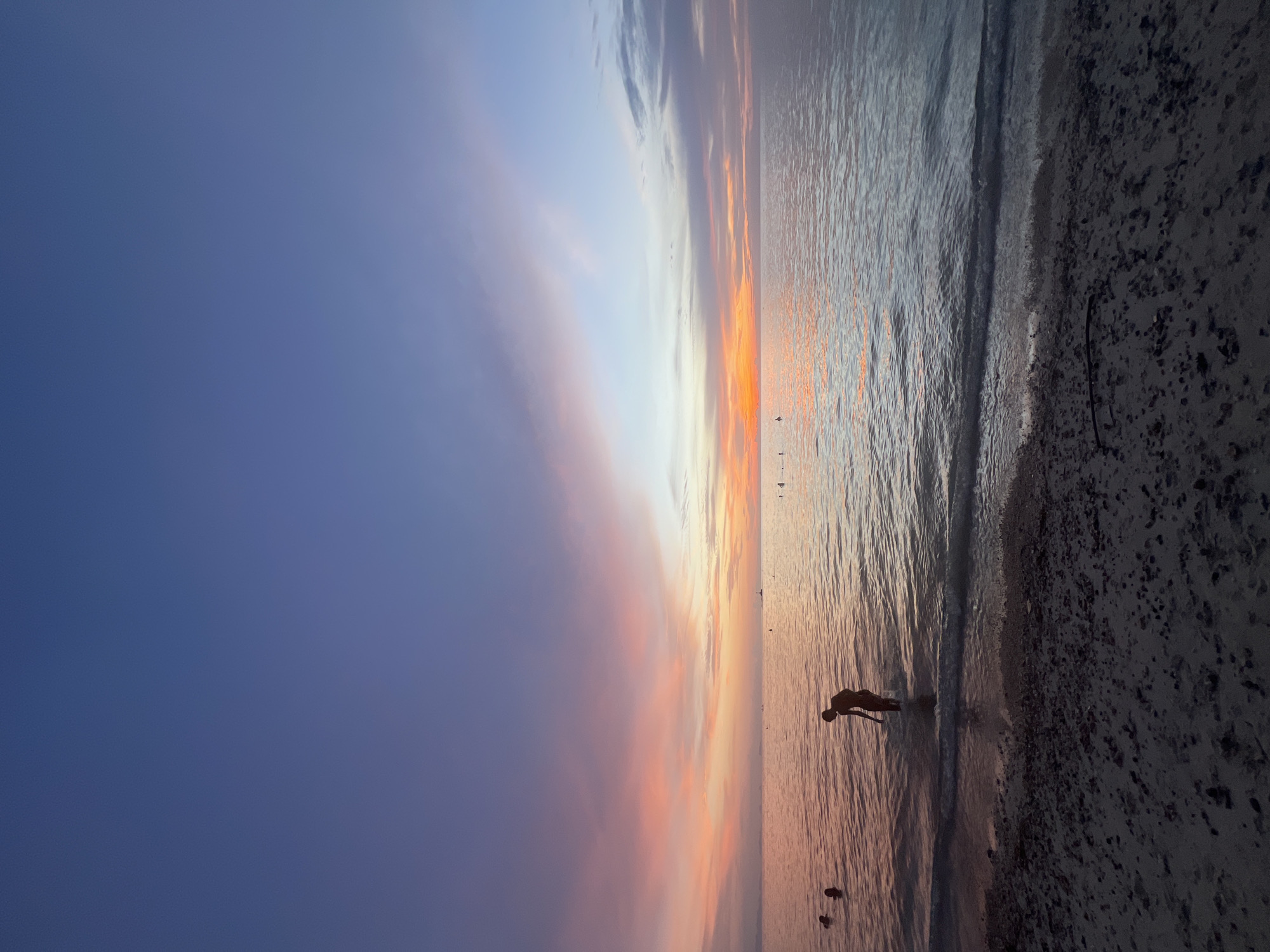 A silhouette of a person stands in shallow water on a beach at sunset. The sky is a gradient of deep blue to pink and orange, with clouds reflecting the sunset colors. The ocean is calm, with gentle waves lapping at the shore. The beach is sandy and scattered with small pebbles. In the distance, several other people are visible in the water and on the horizon, along with a faint lighthouse. The location is likely a beach on Ko Pha-ngan District, Thailand, given the visual characteristics.