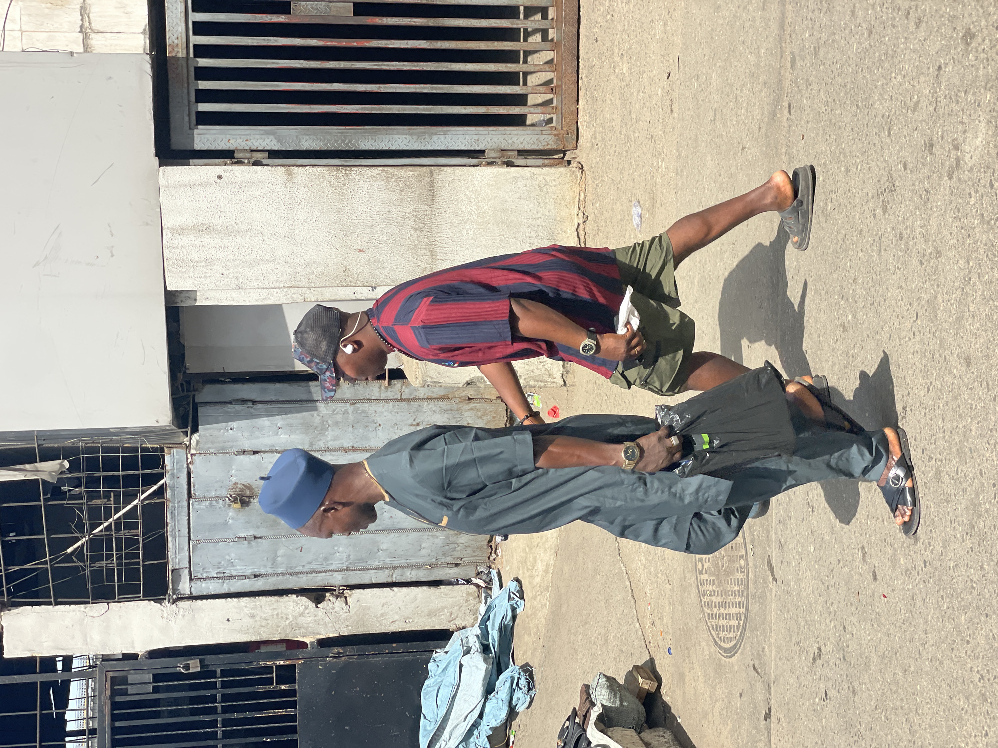 Two men walk side by side on a concrete pavement in Lagos, Nigeria. The man in the foreground wears a dark blue outfit and a matching cap. He carries a black plastic bag. The second man wears a red and blue striped shirt, khaki shorts, and a baseball cap. He has earbuds in his ears. Both men wear sandals. They are walking in front of buildings with weathered, industrial-looking metal doors and barred windows. There is a pile of discarded clothing or fabric on the ground near the building. The day is sunny.