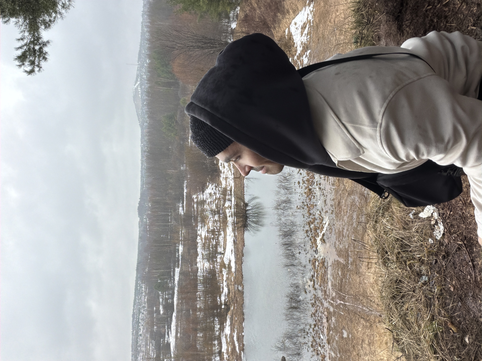 A medium shot depicts a person in profile, dressed in a light-colored sweatshirt with a dark hood, standing at the edge of a bluff. The individual also wears a dark beanie and has a backpack slung over their shoulder. The person is looking downward, appearing to be gazing at a body of water below. Patches of snow are visible around the water's edge and on the nearby land, suggesting a winter or early spring setting. The background includes bare trees and what appears to be a distant urban area possibly the city of Izhevsk, Russia. The sky is overcast, which contributes to the subdued, muted tones of the photograph. The image is rotated 90 degrees counterclockwise.