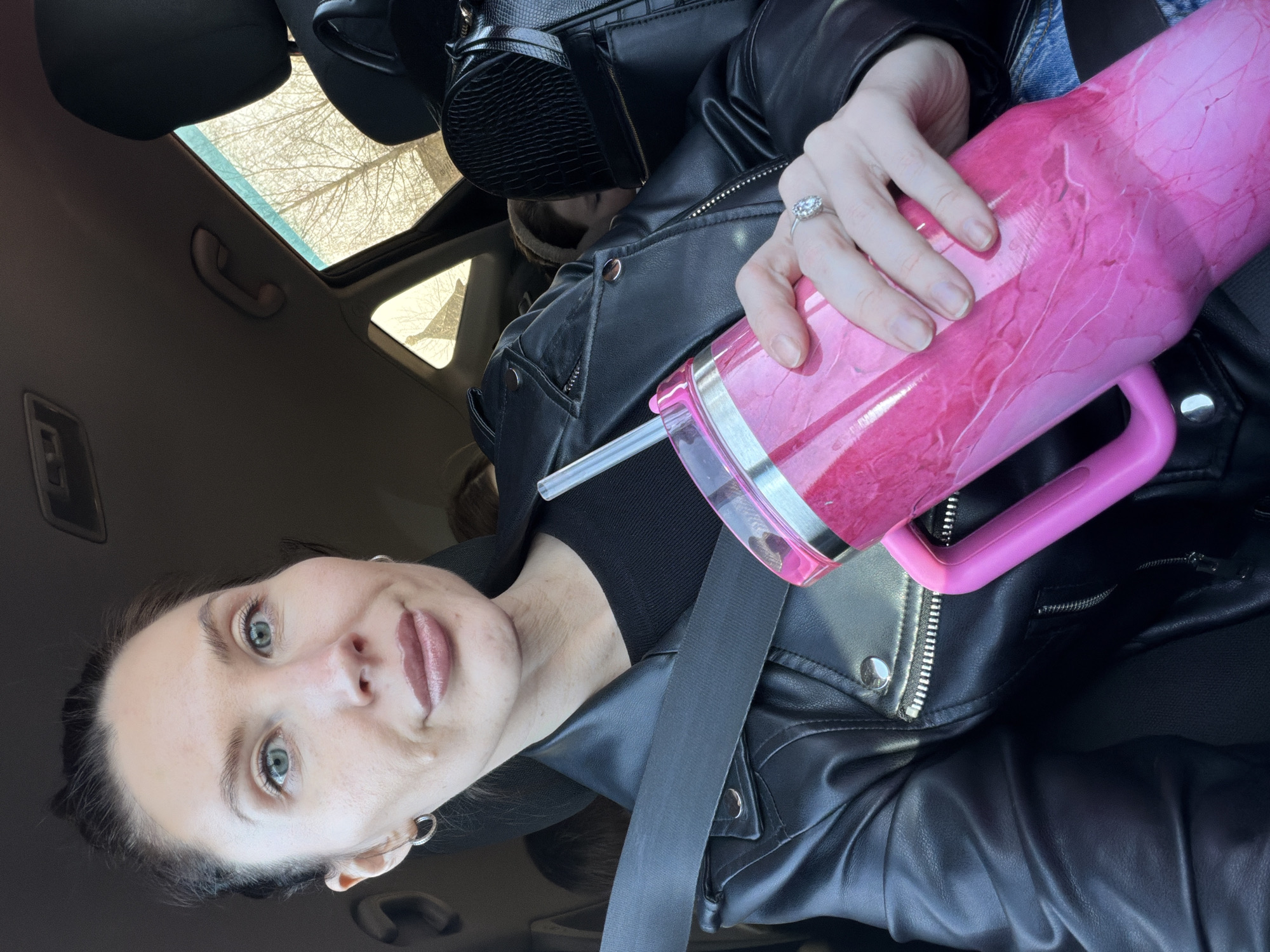 A selfie of a woman with light skin and green eyes, wearing a black leather jacket and a grey seatbelt in what appears to be the driver's seat of a car. She is holding a pink tumbler with a clear lid and a straw in her right hand. The car has a sunroof open, revealing bare tree branches against a bright sky. A black purse is visible. The woman is wearing a silver ring on her ring finger and hoop earrings. The background is a blurred interior of the car. There is no evidence in the image to suggest that the location is Perm, Russia.