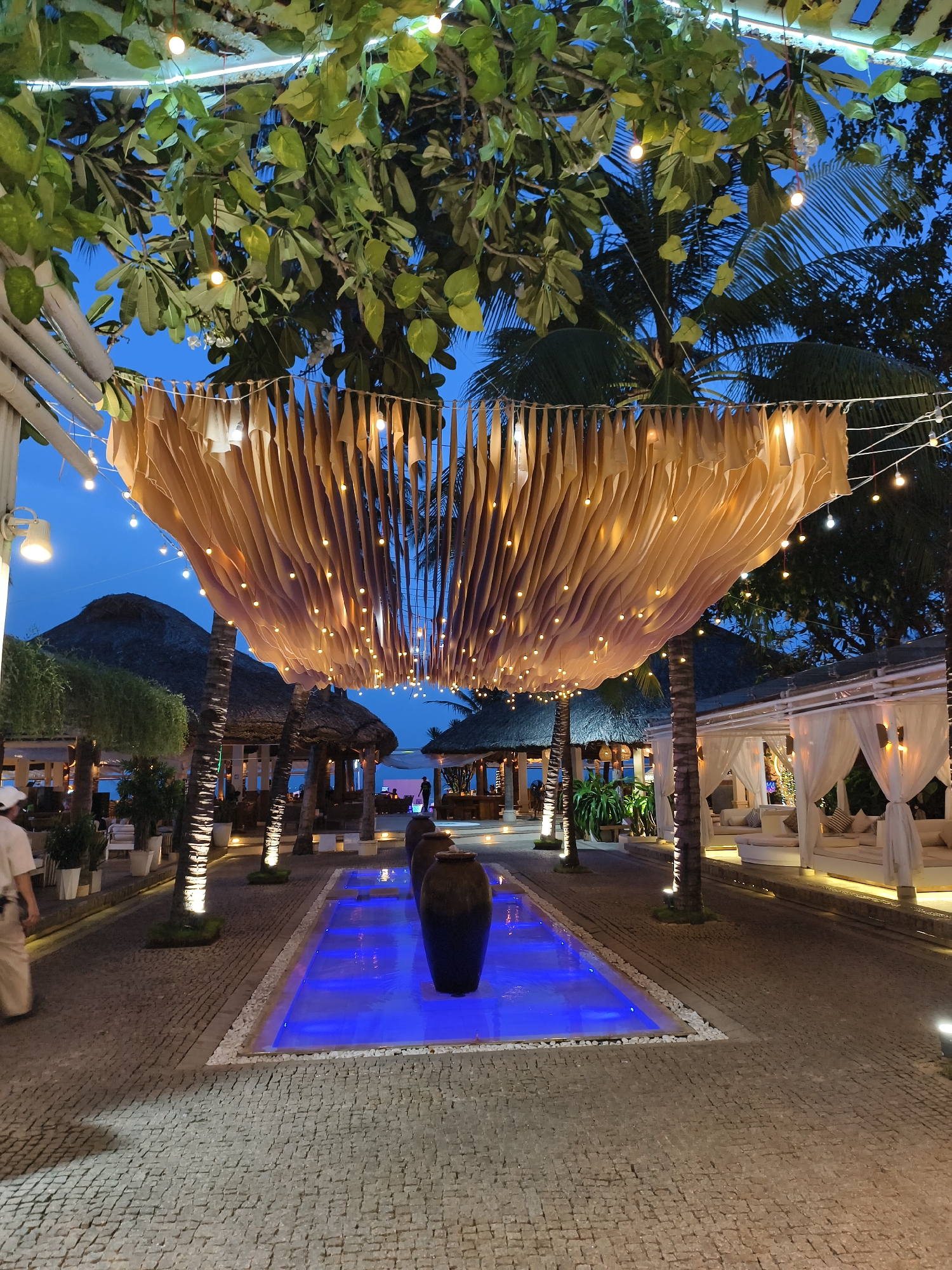 This image depicts an outdoor pathway in Nha Trang Ward, Vietnam, lined with palm trees and draped with a canopy of light-colored fabric adorned with hanging lights. A blue-lit water feature runs down the center of the pathway, accented with dark-colored urns. On the right side, there are lounge areas with white canopies and seating. In the background are buildings with thatched roofs, and the evening sky is visible above. A person can be seen walking in the distance. The ground is paved with cobblestones.