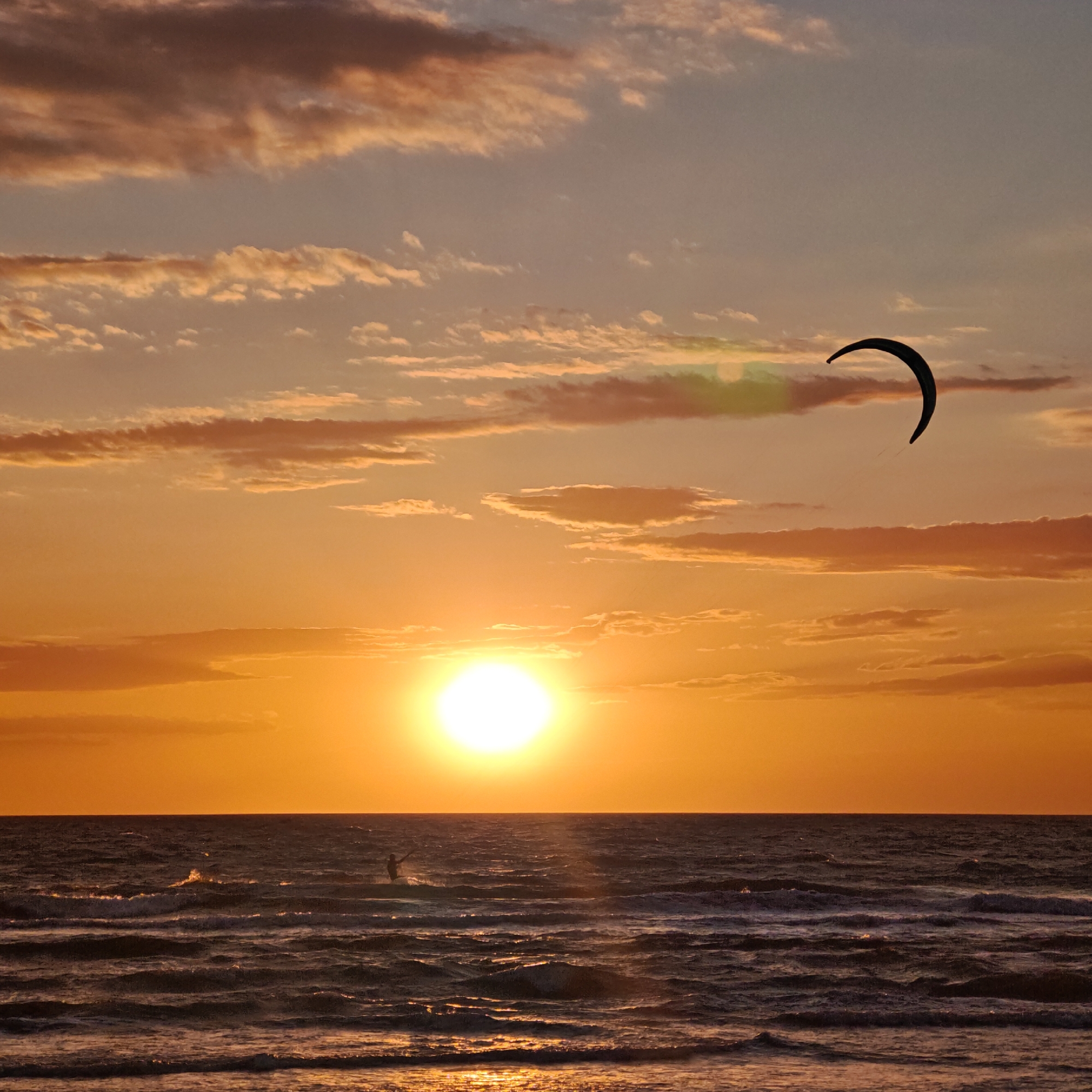 A sunset over the ocean. In the foreground, waves gently roll towards the viewer. A figure, likely a kitesurfer, stands on the water near the horizon. Above, the sun shines brightly, partially obscured by clouds. In the upper right, a kite is visible in the sky. The scene evokes warmth and tranquility. The location in the city of Unknown, Unknown.