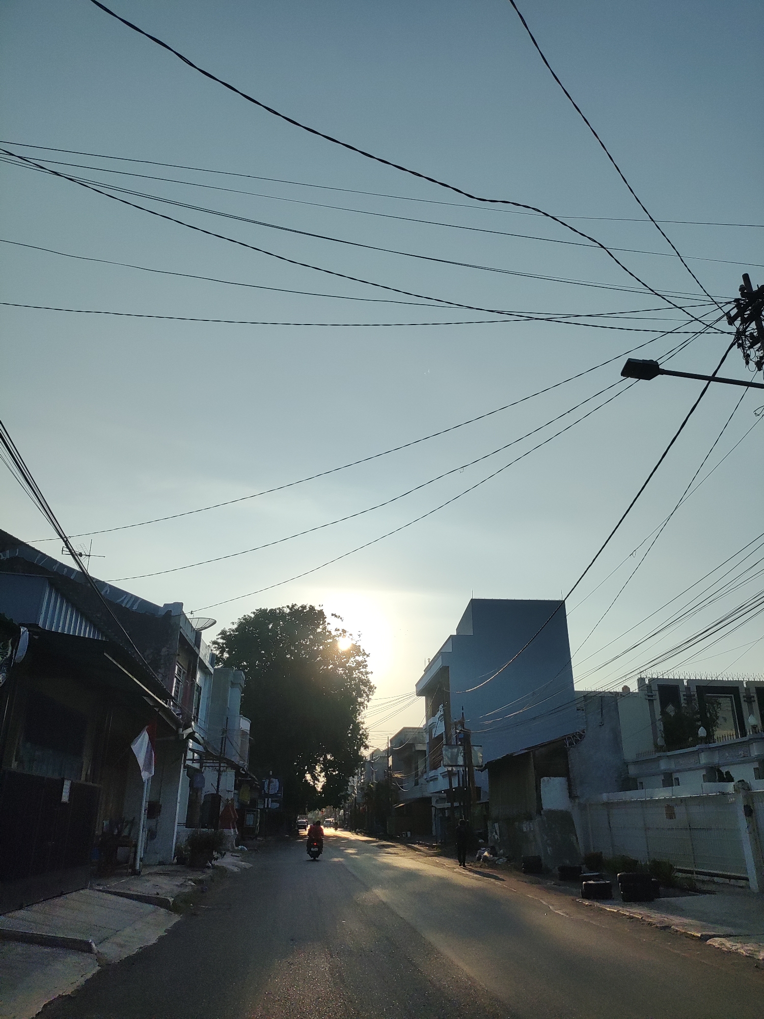The image depicts a street scene in Cirebon, Indonesia, during the late afternoon or early morning, as indicated by the low angle of the sun casting long shadows. The sky is clear and pale blue, crisscrossed by a dense network of utility wires. Buildings line both sides of the asphalt road, with a mix of architectural styles, including some with corrugated metal roofs and others with more modern facades.
On the left side of the road, there are low-rise buildings, one of which displays an Indonesian flag. The other side of the street features a larger, multi-story building with a light blue facade, and further down, a walled property with ornate metalwork. A prominent, large tree with dense foliage stands near the center of the frame, partially obscuring the sun.
A person on a scooter, with their back to the camera, is traveling away from the viewer down the road. Another individual, appearing as a dark silhouette, is walking on the sidewalk on the right side of the street. The road surface is mostly clear, with some scattered debris and what appear to be discarded tires placed along the right edge of the road. A street lamp is visible on the right, integrated with a utility pole laden with wires. The overall impression is of a typical urban street in a Southeast Asian city, bathed in soft sunlight.