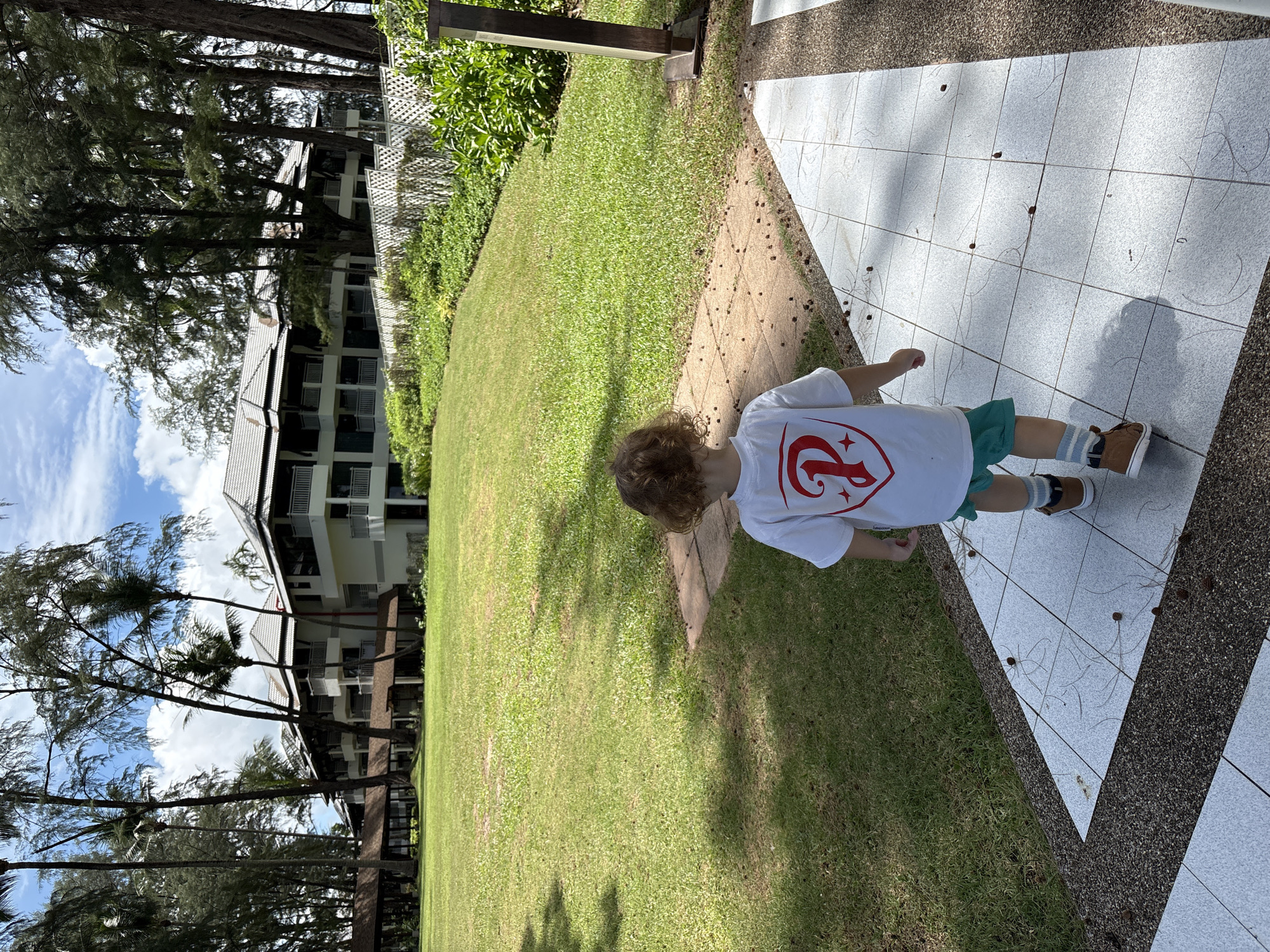 In the city of Choeng Thale, Thailand, a young child with curly brown hair walks on a tiled pathway. The child is wearing a white t-shirt with a red emblem, green shorts, and brown shoes with blue and white striped socks. The pathway is surrounded by a manicured green lawn, and in the background, a large, multi-story building with white walls and dark roofs is visible. Tall trees, some with palm fronds, line the property, and a white lattice fence separates the lawn from dense foliage. The sky is partly cloudy.