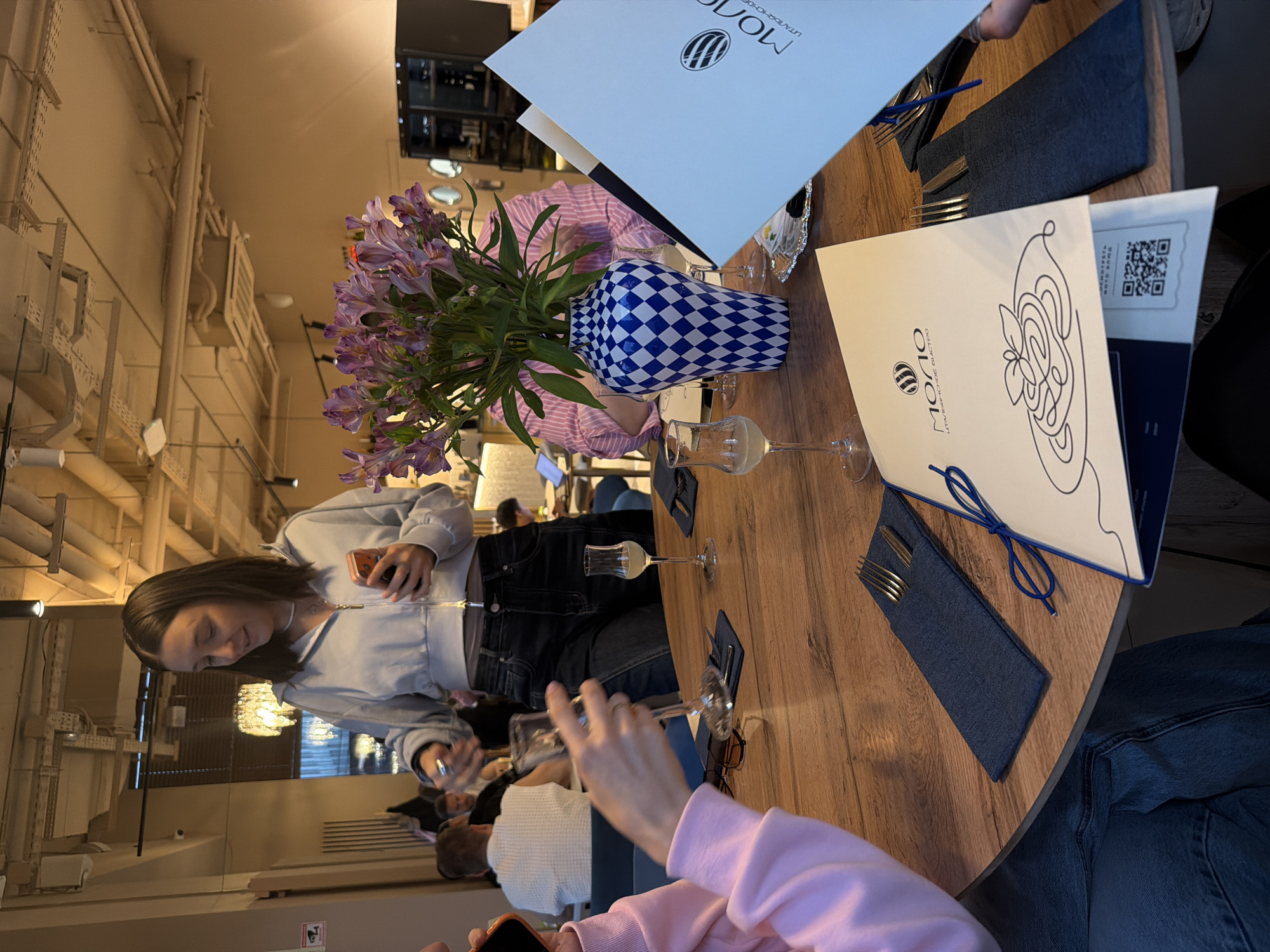 The image shows a restaurant interior with a round wooden table set for dining. On the table, there is a blue and white patterned vase holding a bouquet of purple and pink Alstroemeria flowers. Next to the vase, there are several wine glasses, menus with the logo "MOIIO Italian Bistro", and cutlery sets in denim-like pouches. People are seated around the table, including a woman in a light blue zip-up jacket who is looking at her phone, and a person in a pink sweater holding a smartphone. The scene suggests a social gathering or a meal taking place at the restaurant. The city and location are unspecified.