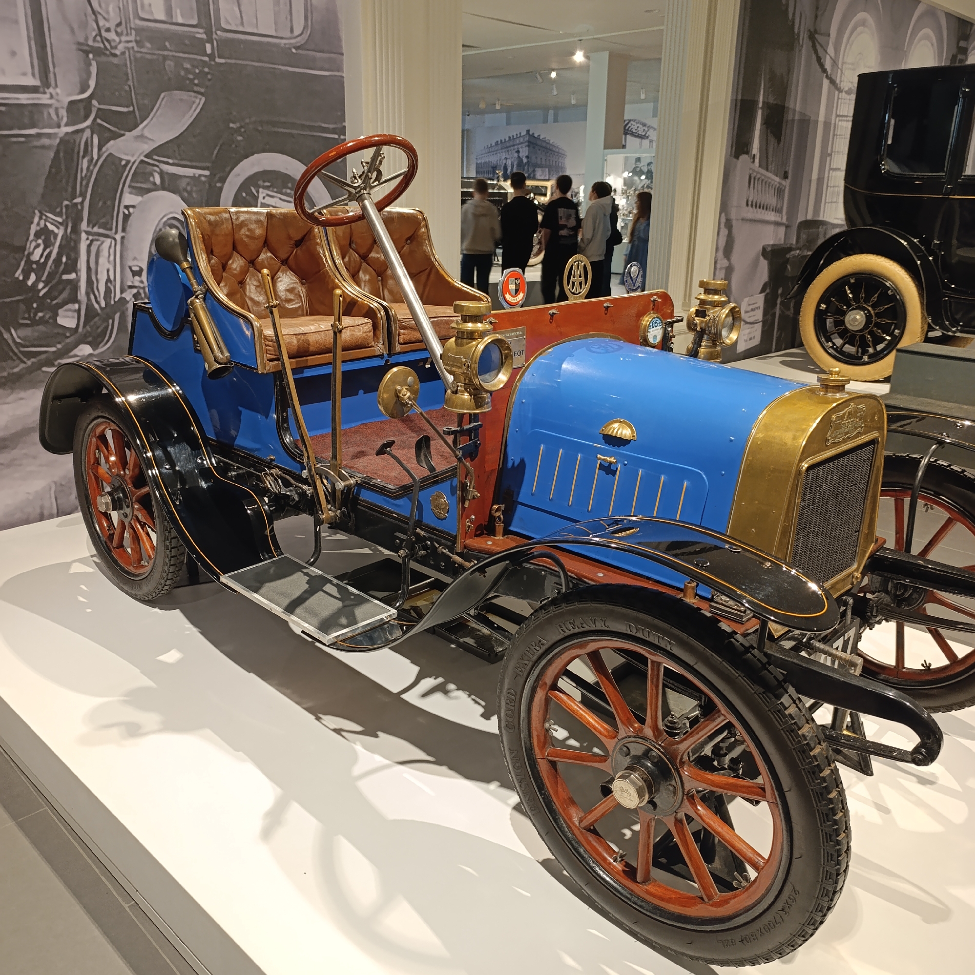 A blue and brown antique car is displayed on a white platform. The car has leather seats, wooden wheels, and brass detailing. In the background, there are other cars and a group of people. The scene appears to be a museum or exhibition. A sign in the background, not fully legible, indicates that the image was taken at the Motor Museum in Verkhnyaya Pyshma, Russia.